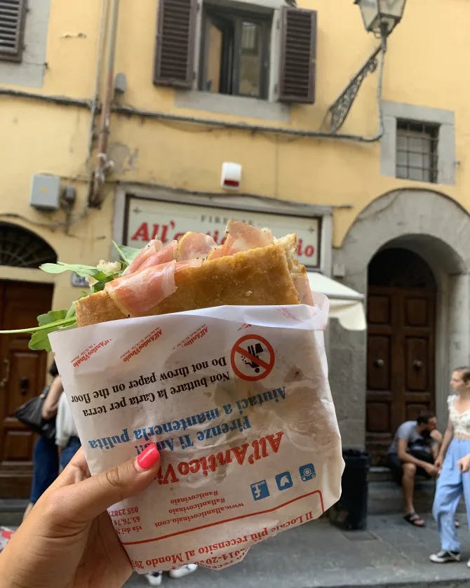 Photo of a someone holding up a focaccia sandwich in front of an old, yellow house