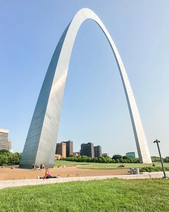 Travel advisor doing the splits on a wall by grass in front of a very large steel arch structure on a sunny day.