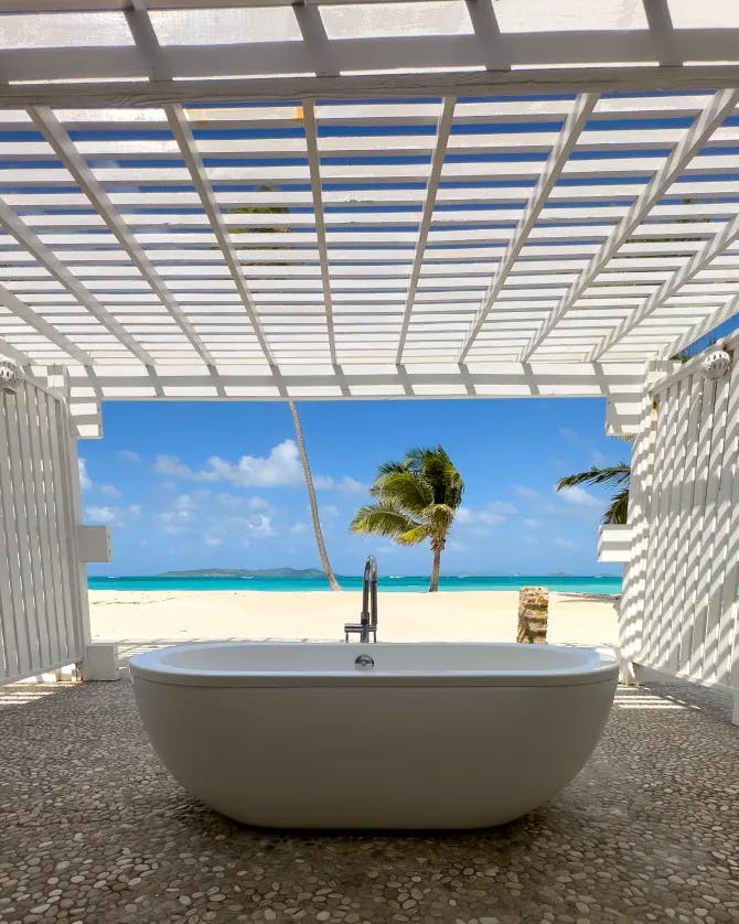 A white bath tub on a stone floor with a beach, palm tree and ocean view in the background