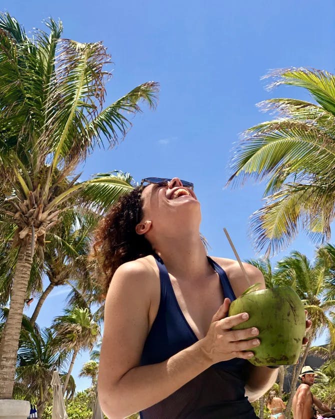 Travel advisor Alexandra looking up at the sky and smiling, holding a green coconut, with palm trees in view