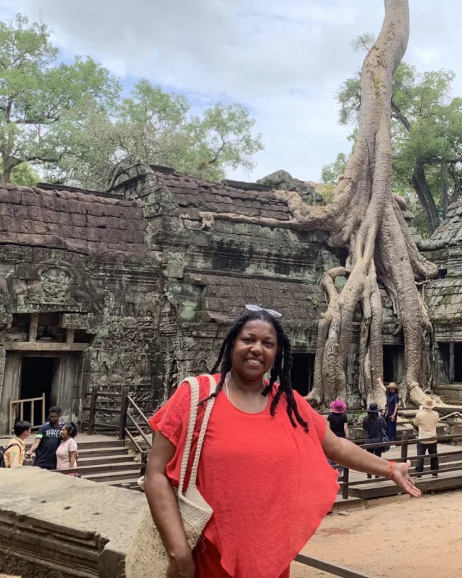 Lynore posing in a red top outside in front of an old stone temple and trees with left arm outstretched. 