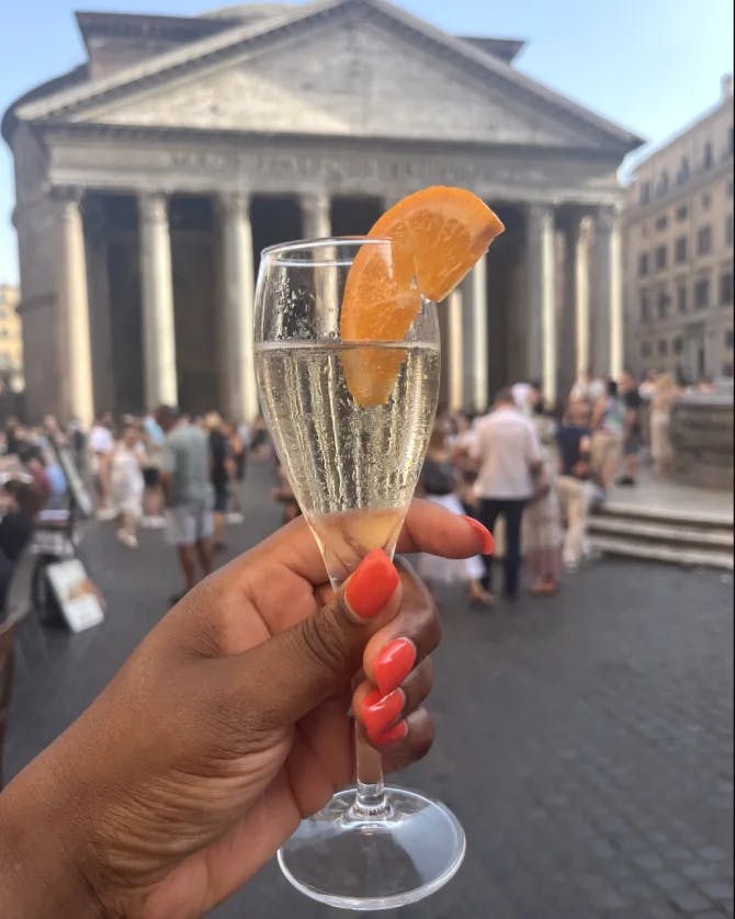 A person with pink nail polish on holding a champagne flute with an orange slice on it. There are people walking around a historic square in the background. 