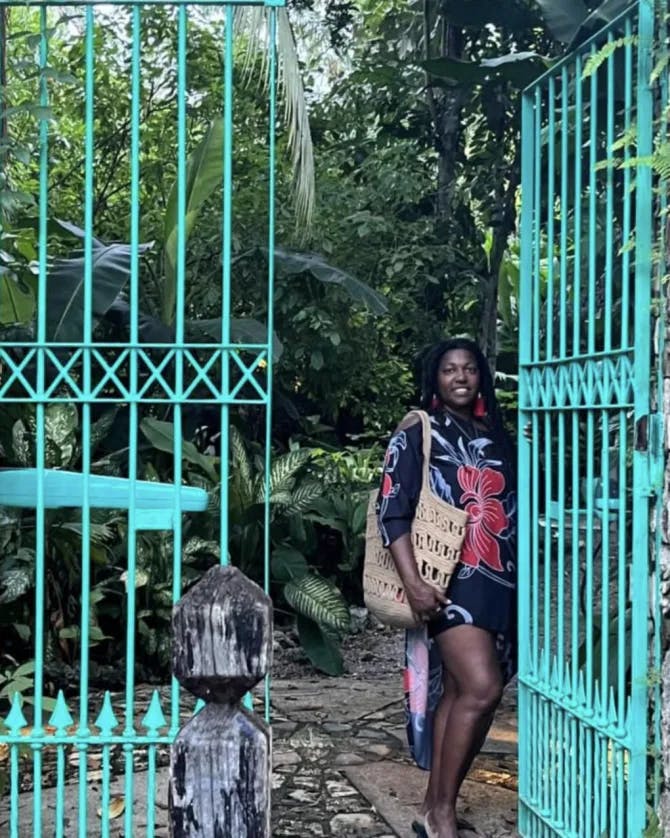 Lynore posing in front of a turquoise gate and trees while wearing colorful clothing. 