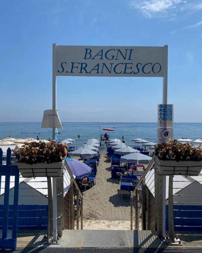 Chairs on a beach covered with blue umbrellas at the bottom of stairs with a banner that says "Bagni S. Francesco."
