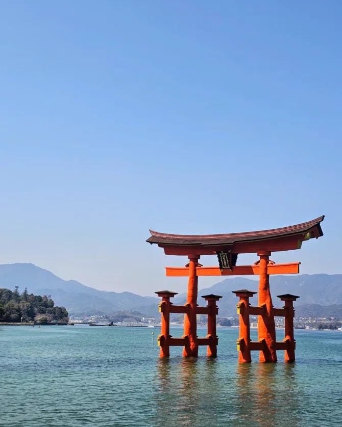 View of iconic "floating" torii gate standing in clear water with mountains in the background on a clear day