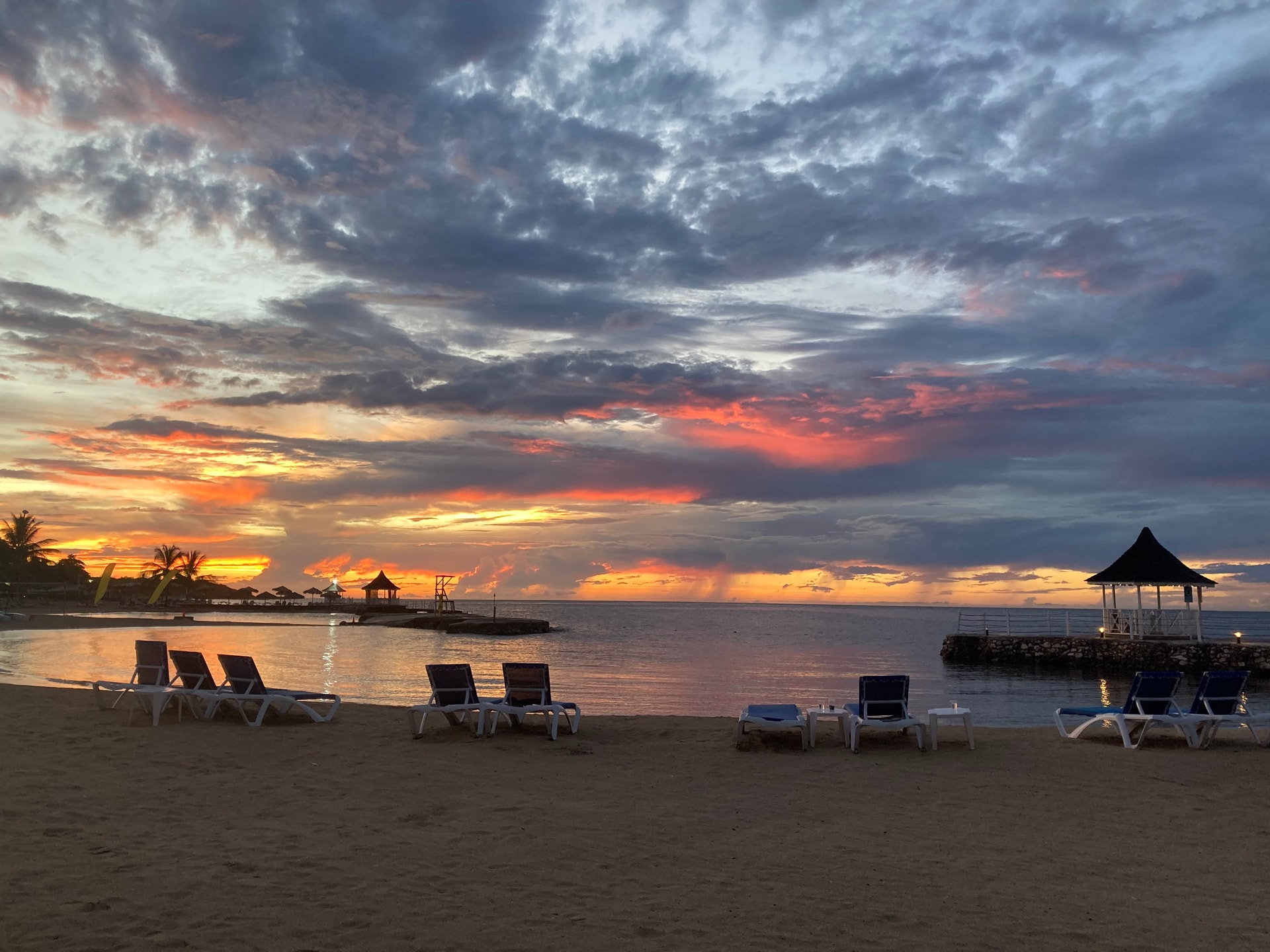 Sunset on the beach with a polychrome sky, chez lounge chairs, and dispersed gazebos. 