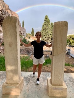 Travel advisor posing in between two marble columns with a rainbow in the sky in the background.