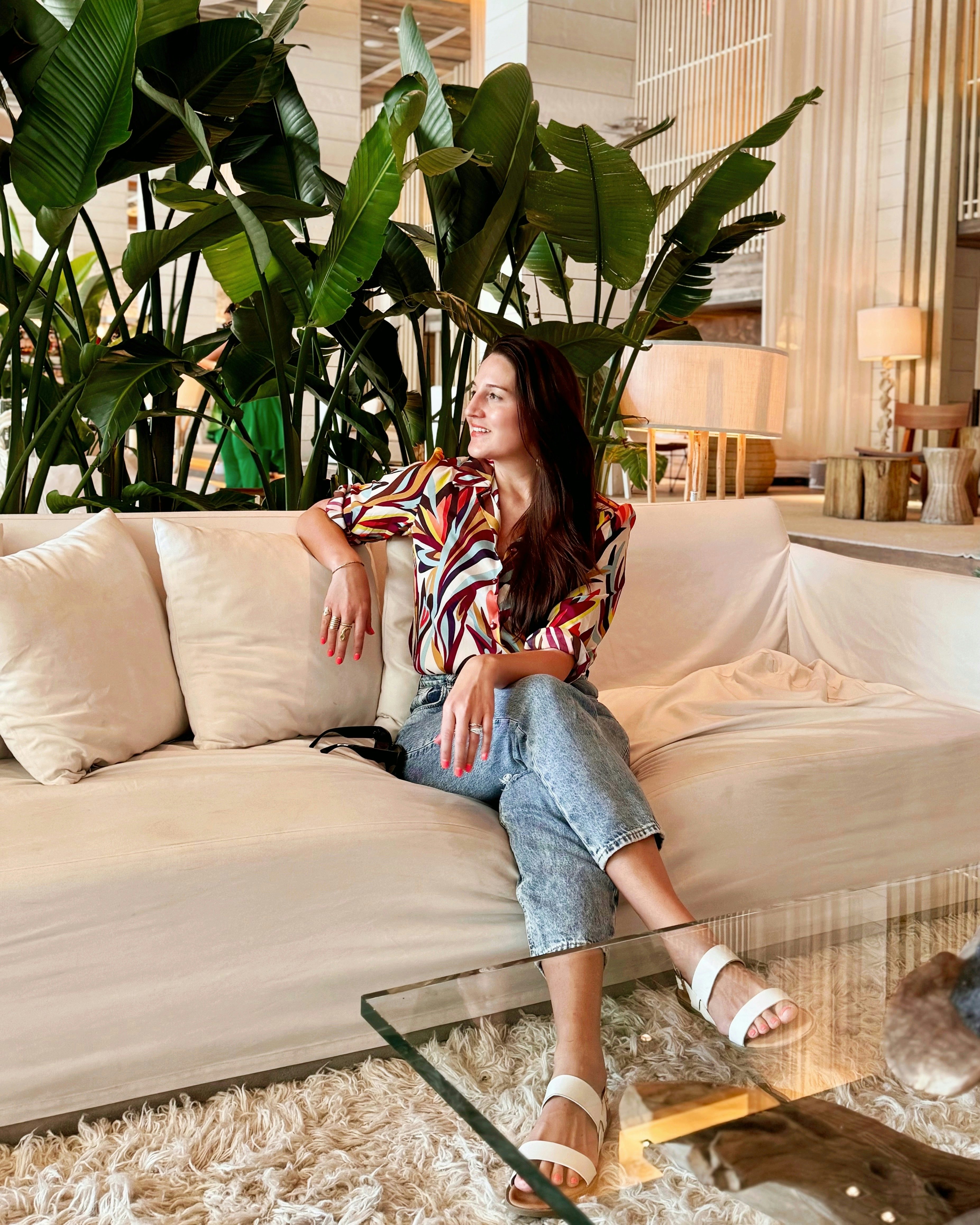 Lauren in jeans with her legs crossed on a beige couch indoors in front of large plants