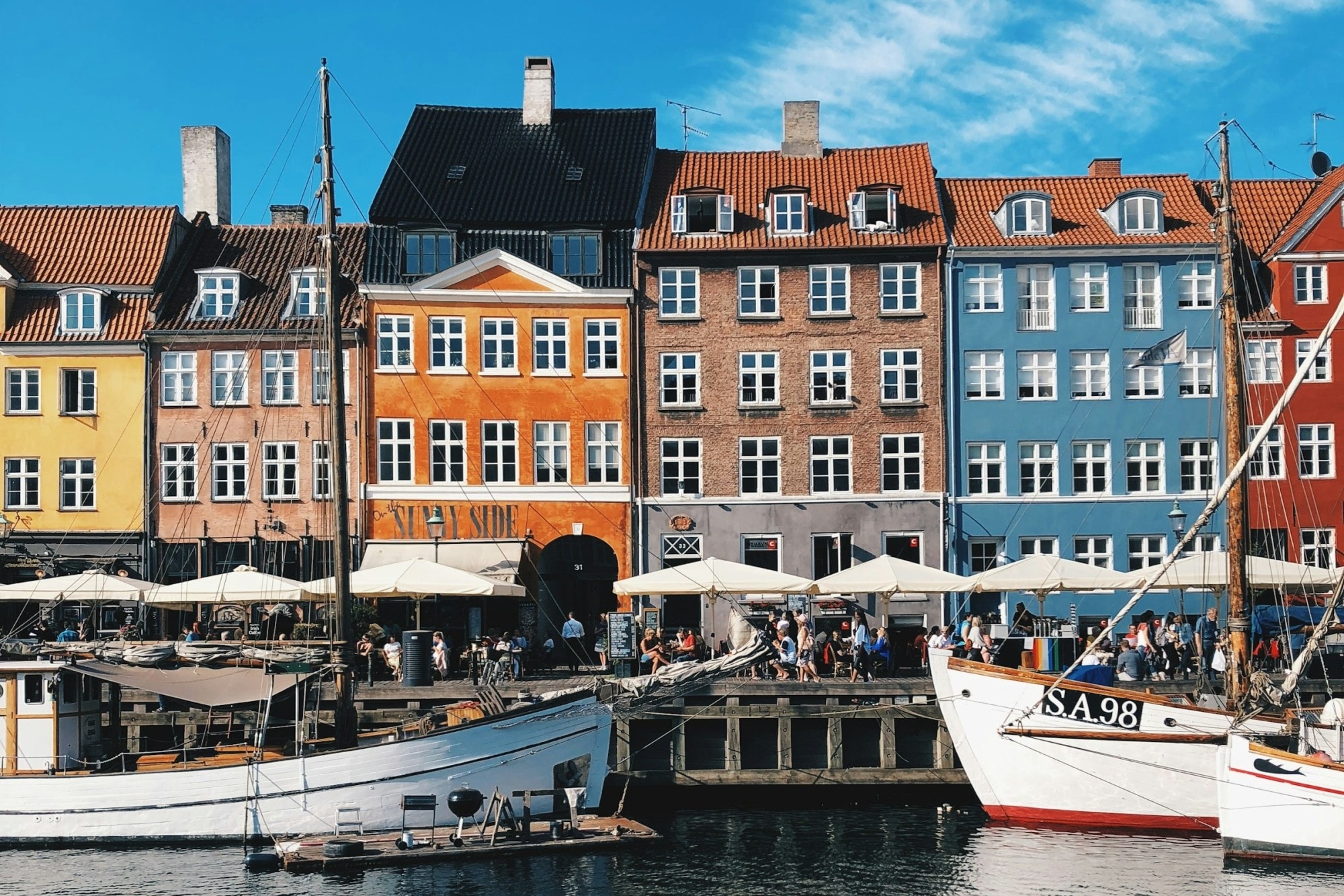 Sailboats dock next to a busy thoroughfare that's lined with colorful, apartment style housing blocks