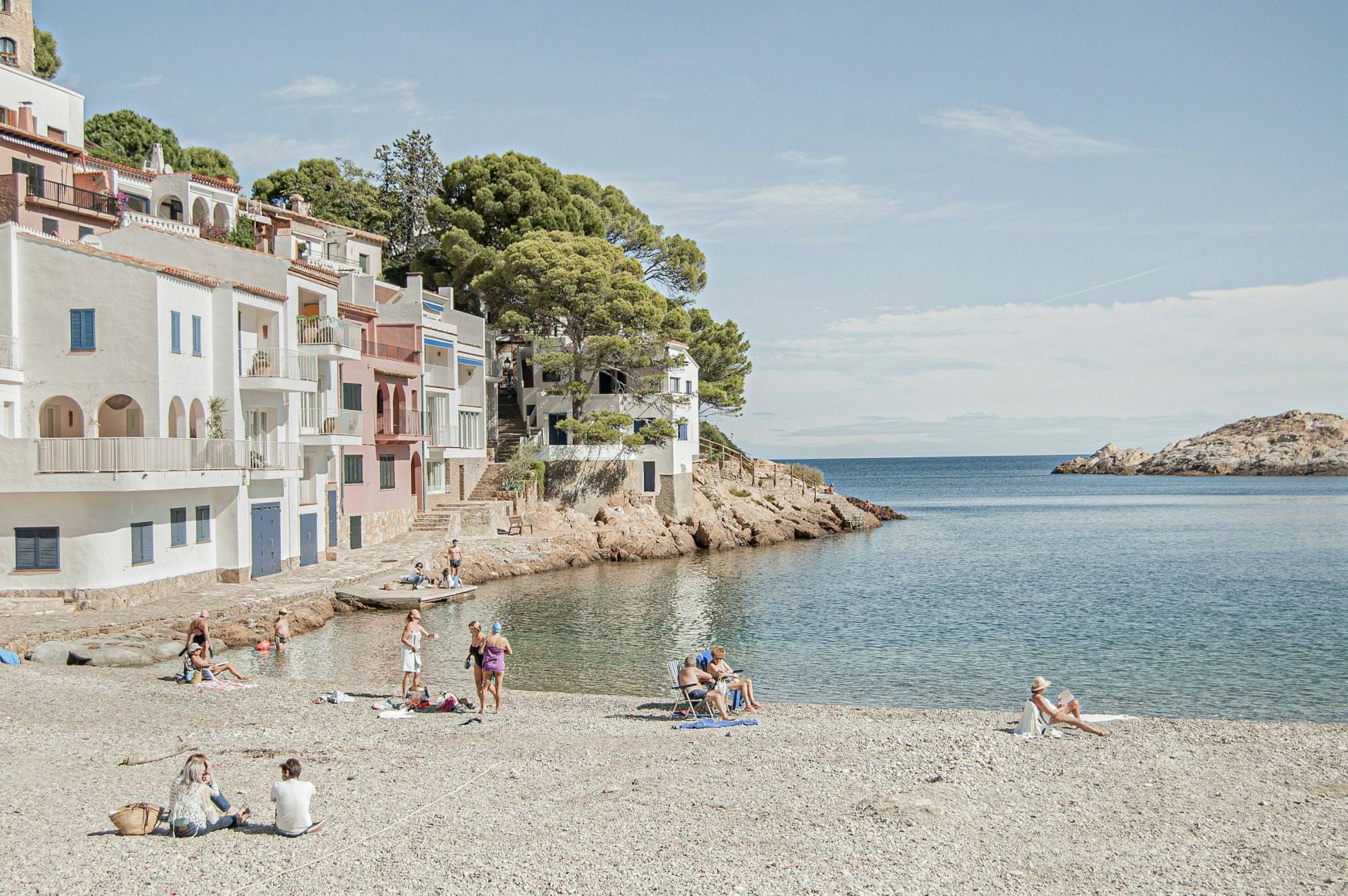 A handful of beachgoers relax on the sand, looking out over calm waters and flanked by historic structrures