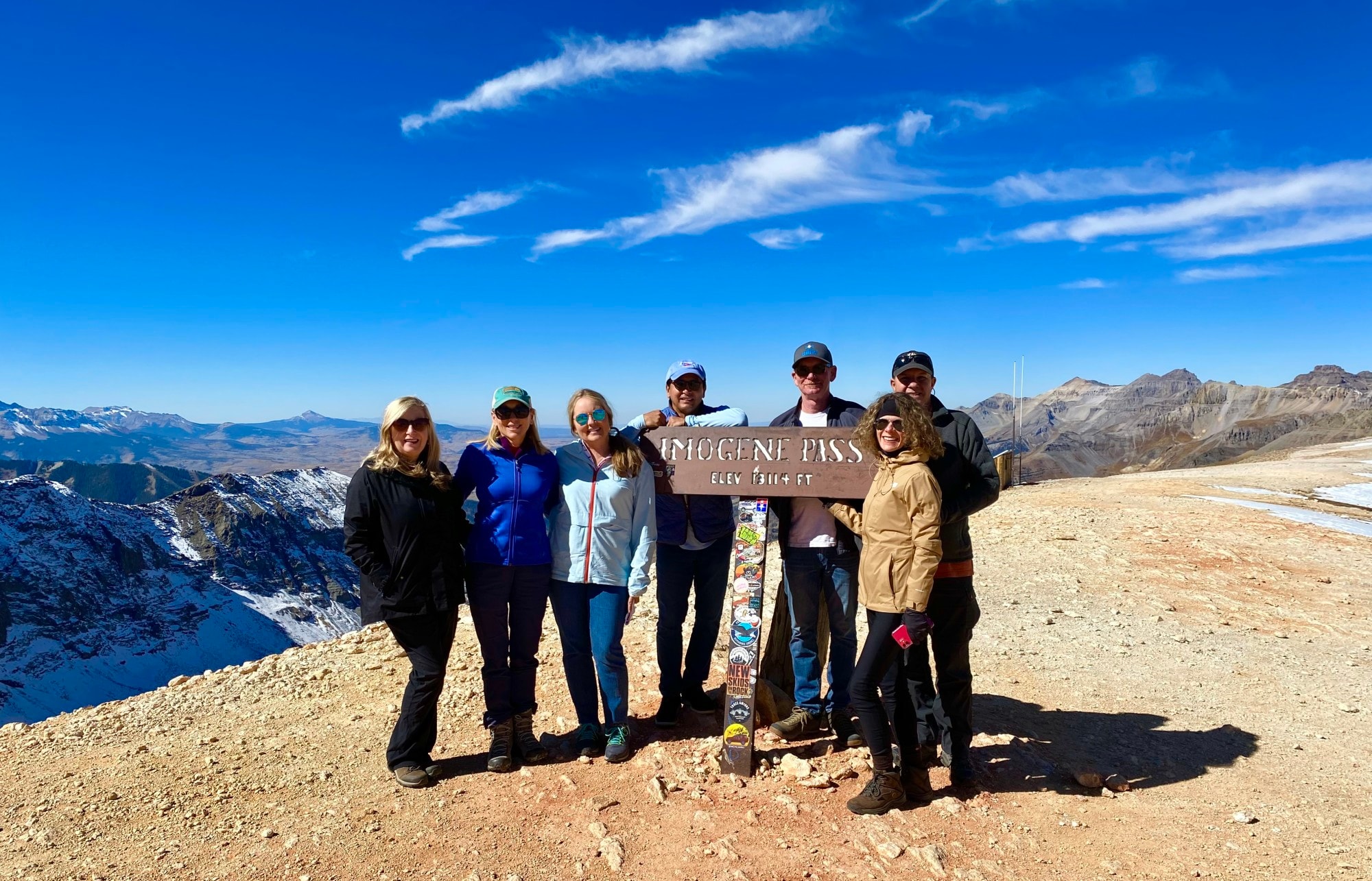 A group of people standing with board saying Imogene pass with snow covered mountains at the back.