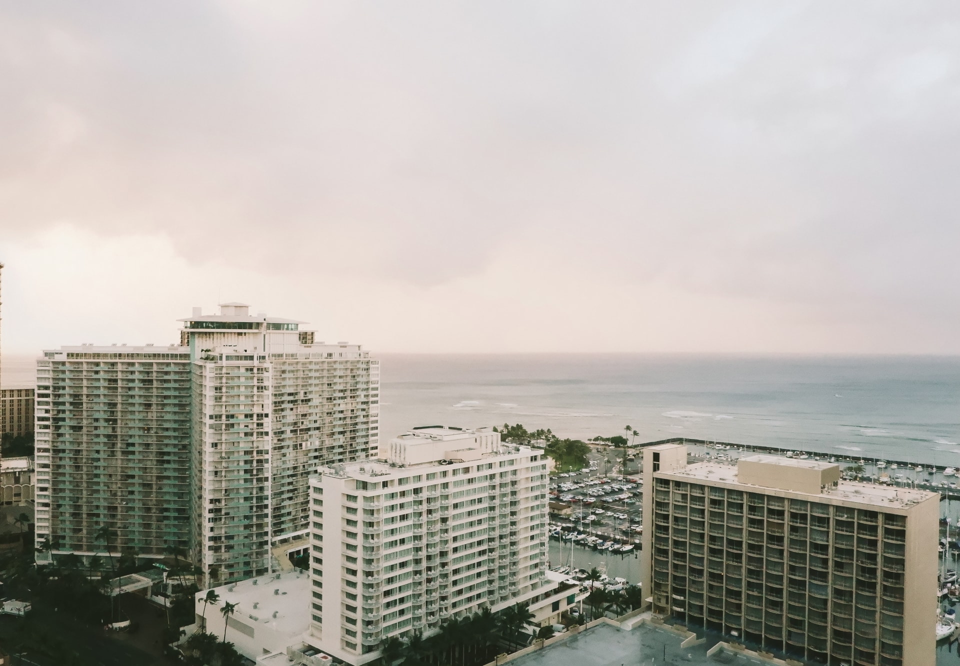 Three white and tan sky rise buildings surrounded by trees, boats and water 