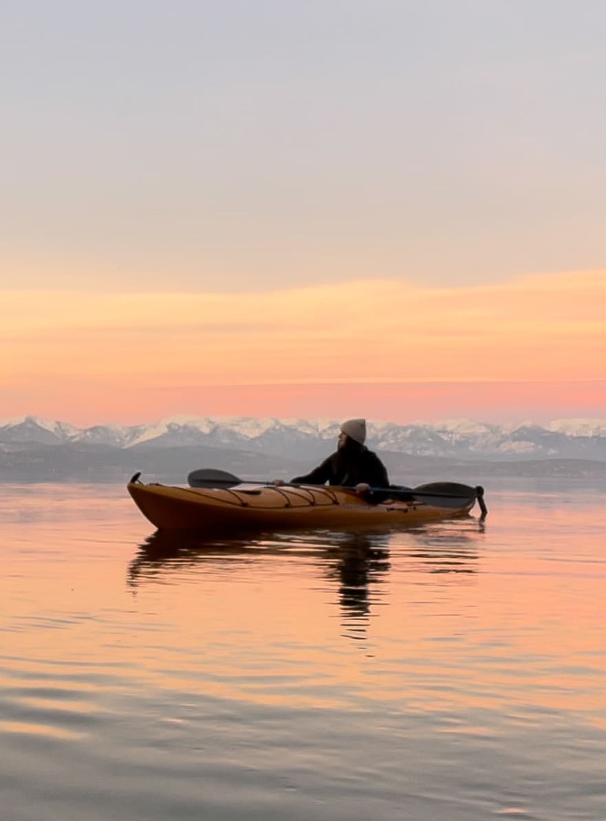 A woman sitting on a canoe with a beautiful light pink sunset reflecting onto the water with mountains in the background. 