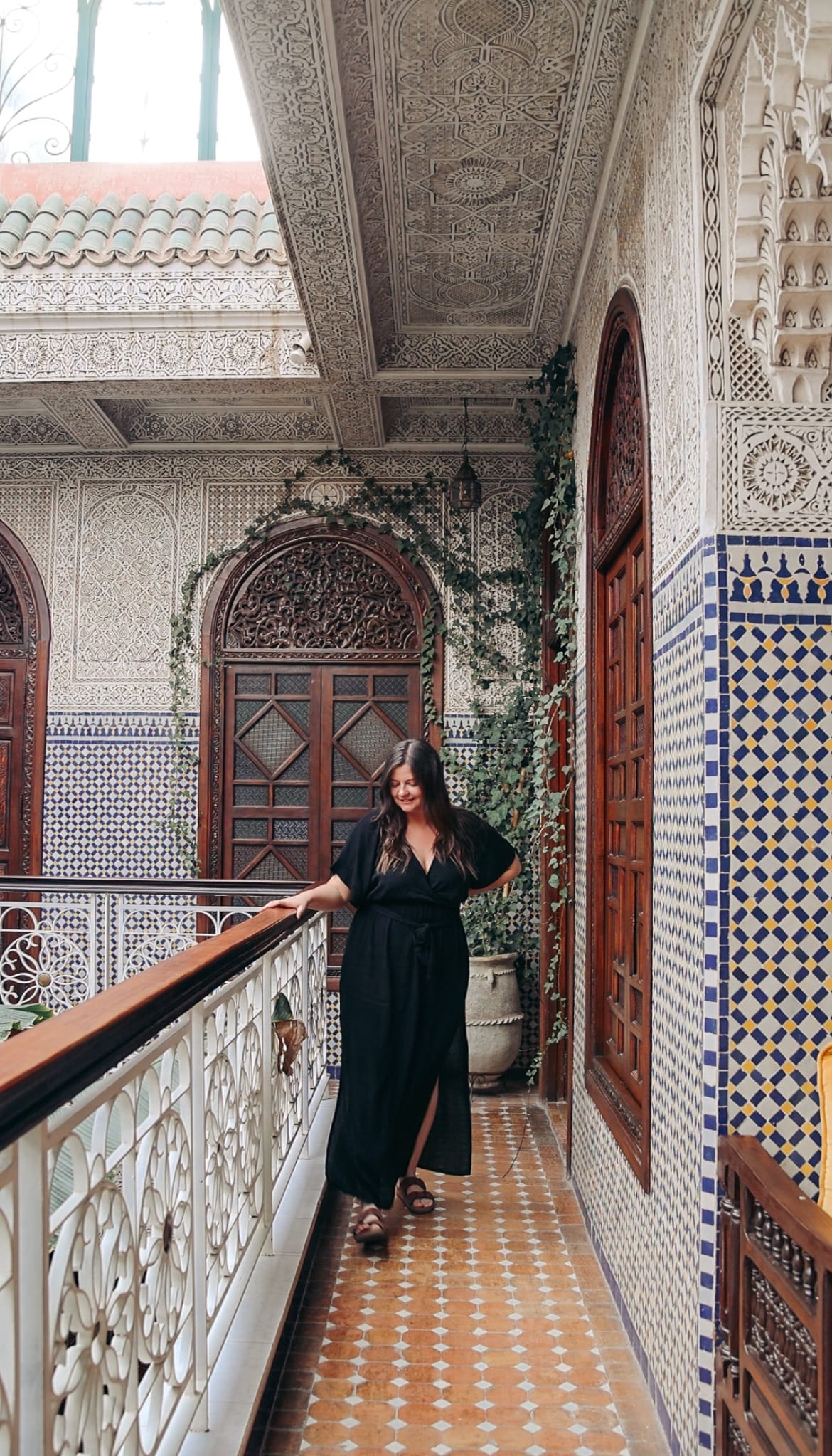 A woman posing in a black dress on a terrace with beautiful mosaic tiling surrounding her and vines on a wall near a wooden arched door in the background. 