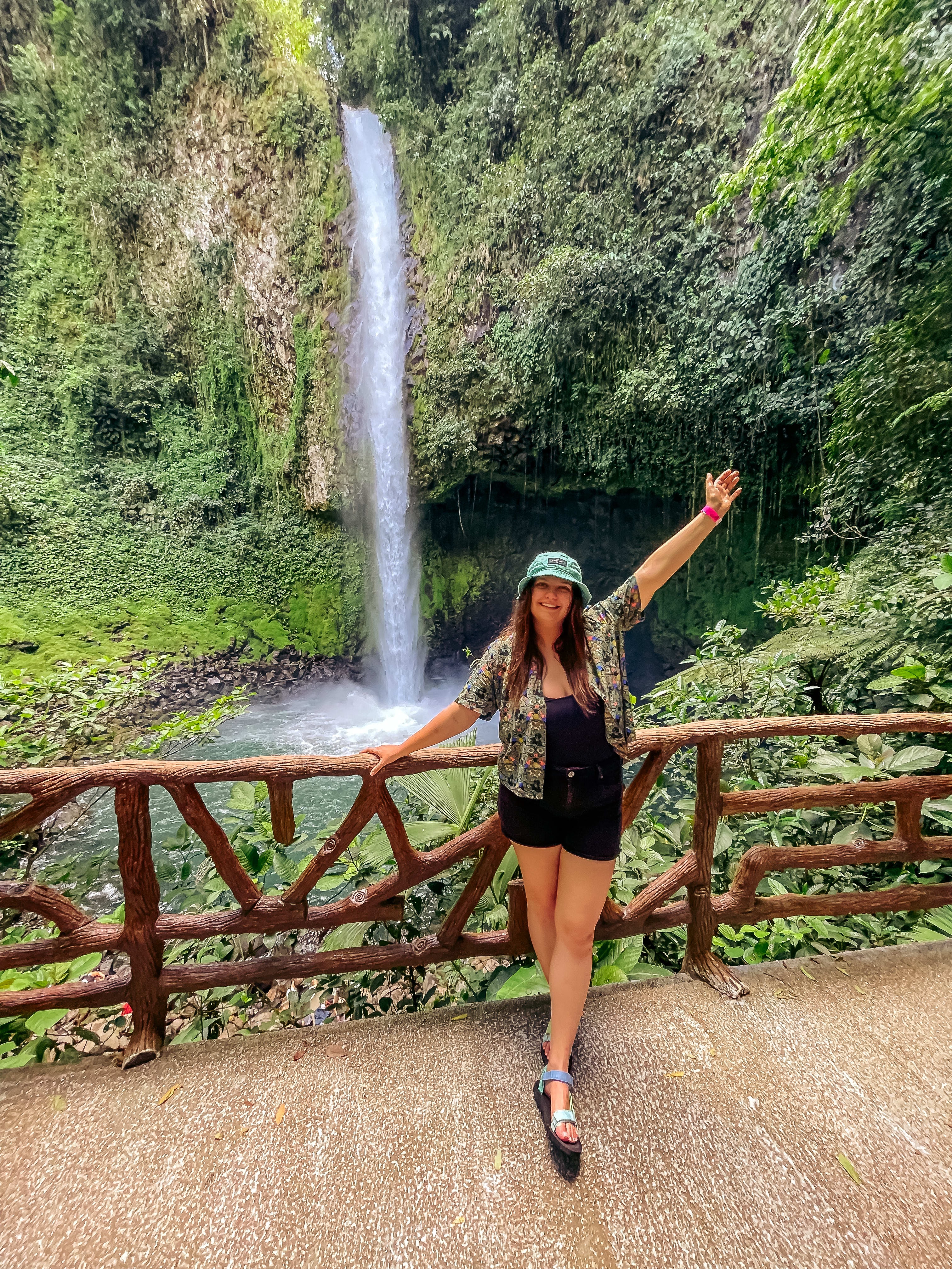 A woman posing with her arm outstretched above her head in front of a view of lush foliage and a waterfall. Her other hand is resting on a wooden fence.  