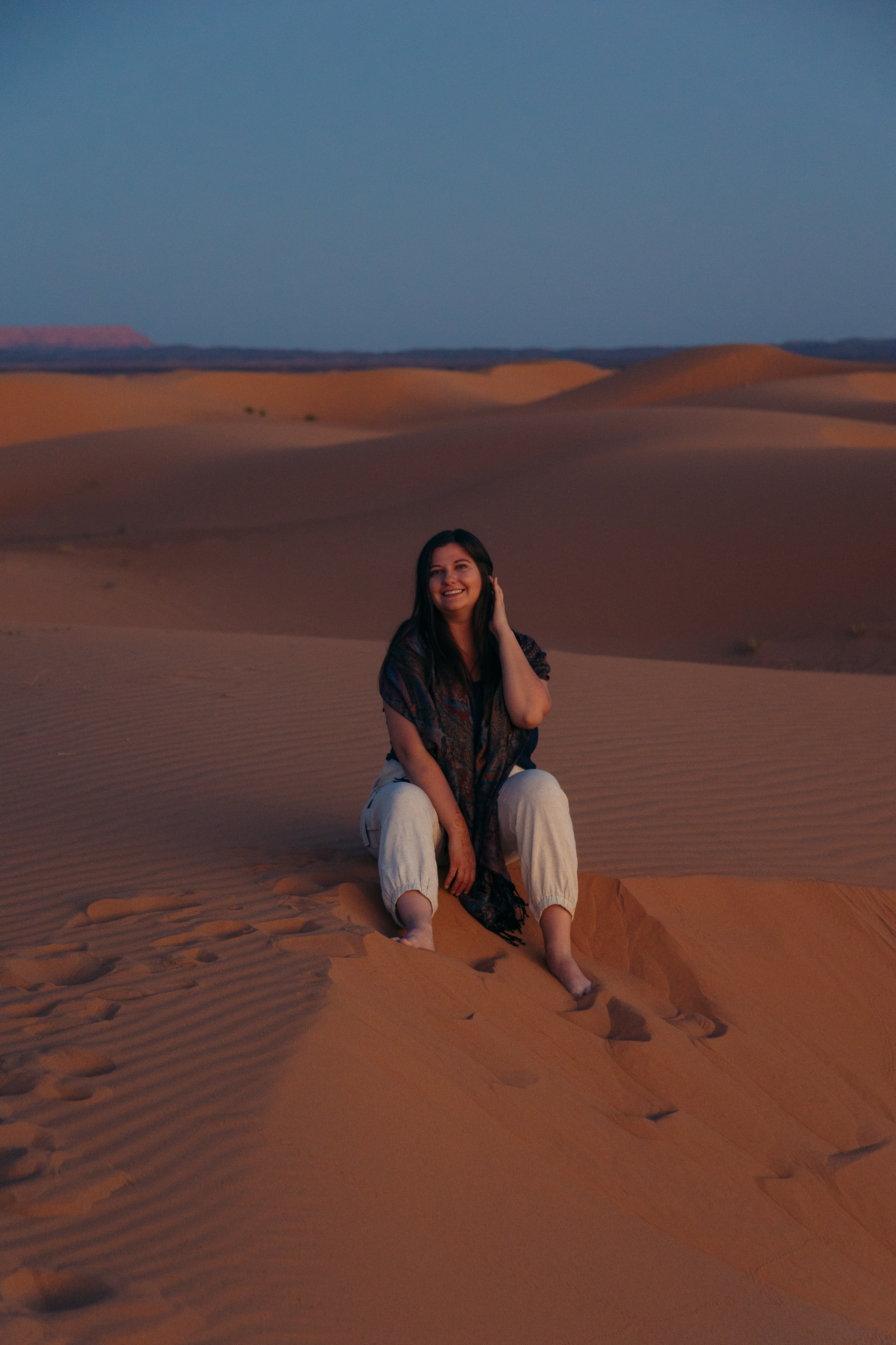 A woman posing for a photo while seated atop of large sand dunes in the desert. She is wearing white pants and a dark top. 