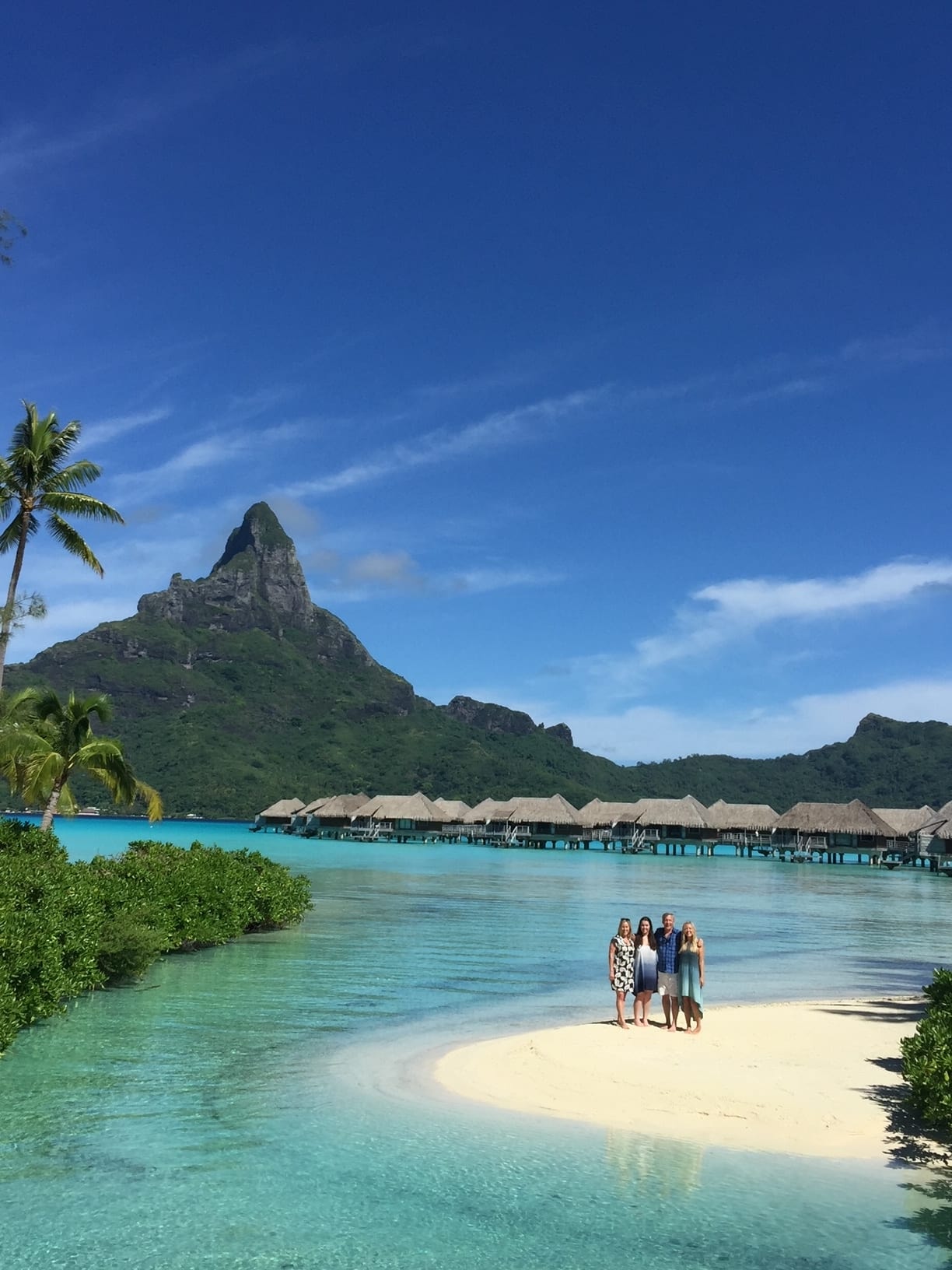 A picture of four people standing on a small sandy inlet surrounded by turquoise water with overwater bungalows in the background