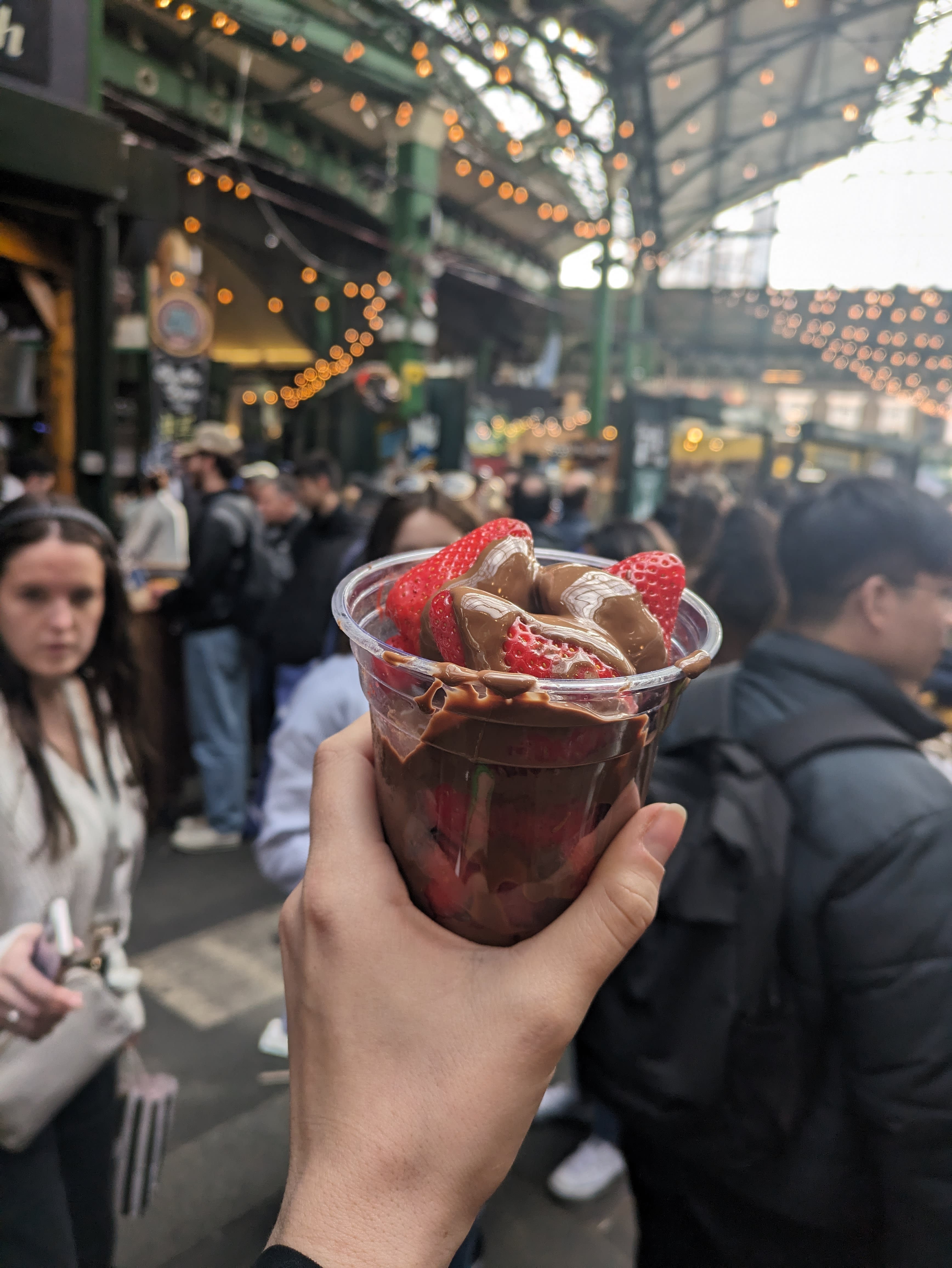 Someone holding up a cup filled with fresh strawberries covered in Nutella in a busy market. 