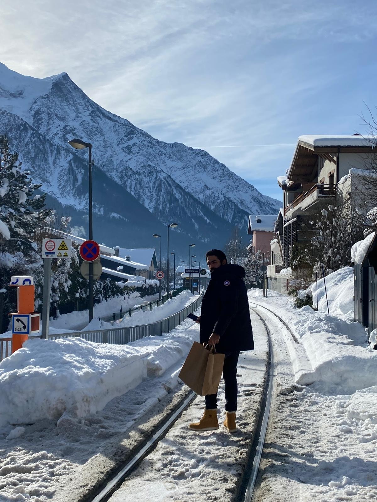 View of Michel wearing winter clothing and standing on a railway track with snowy mountains in the distance