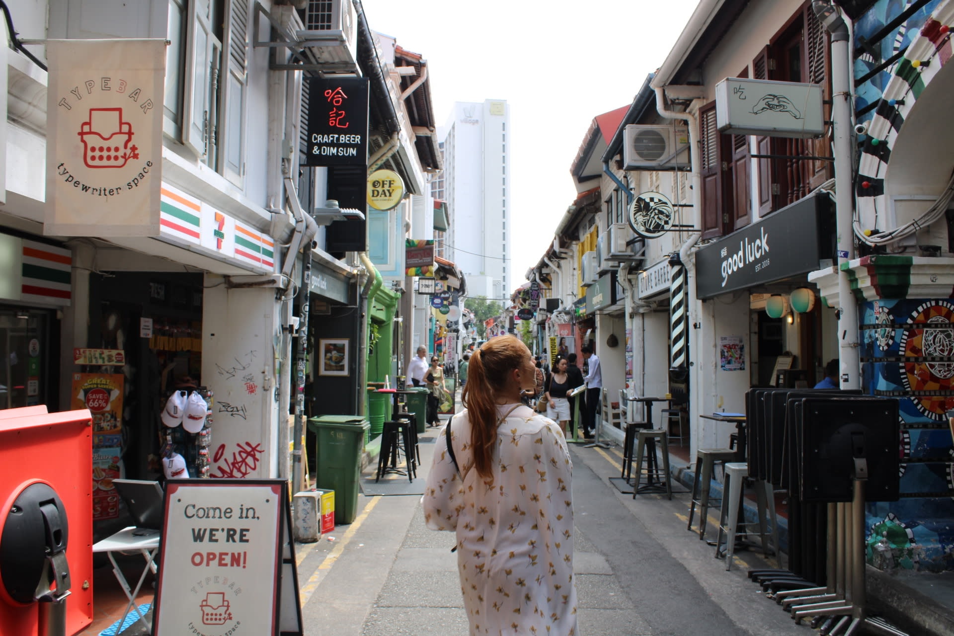 Isabel in a white shawl walking along a street with many buildings and signs on a cloudy day