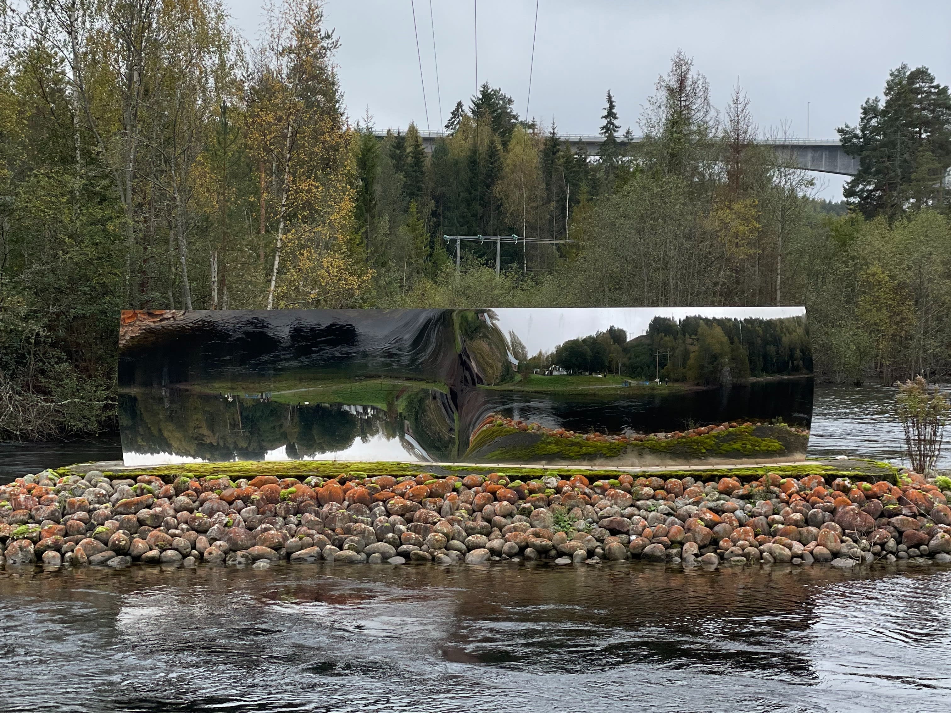 An outdoor mirrored sculpture reflecting greenery and water on a cloudy day.