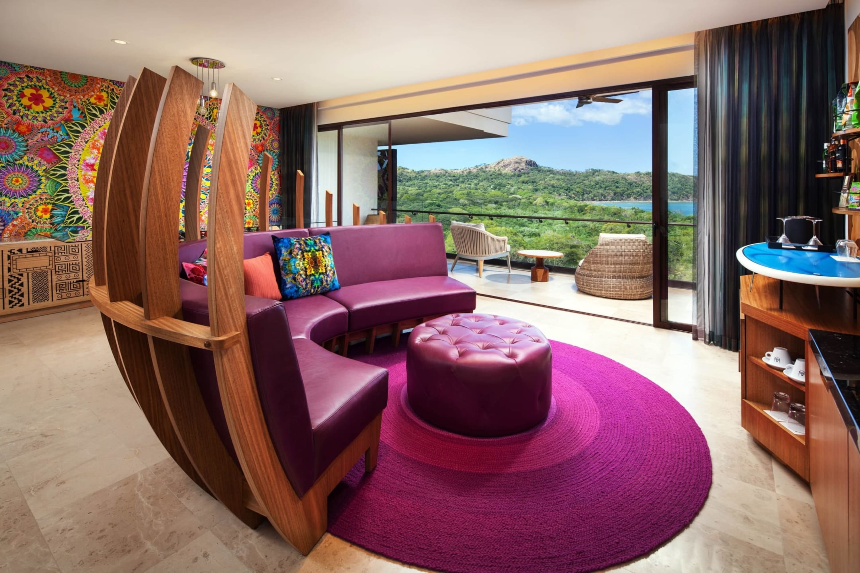 Bold purple and interesting furnishings in a massive living room that opens up to private balcony overlooking Costa Rican jungle and shoreline. A vibrantly colorful mural exists on the opposing wall