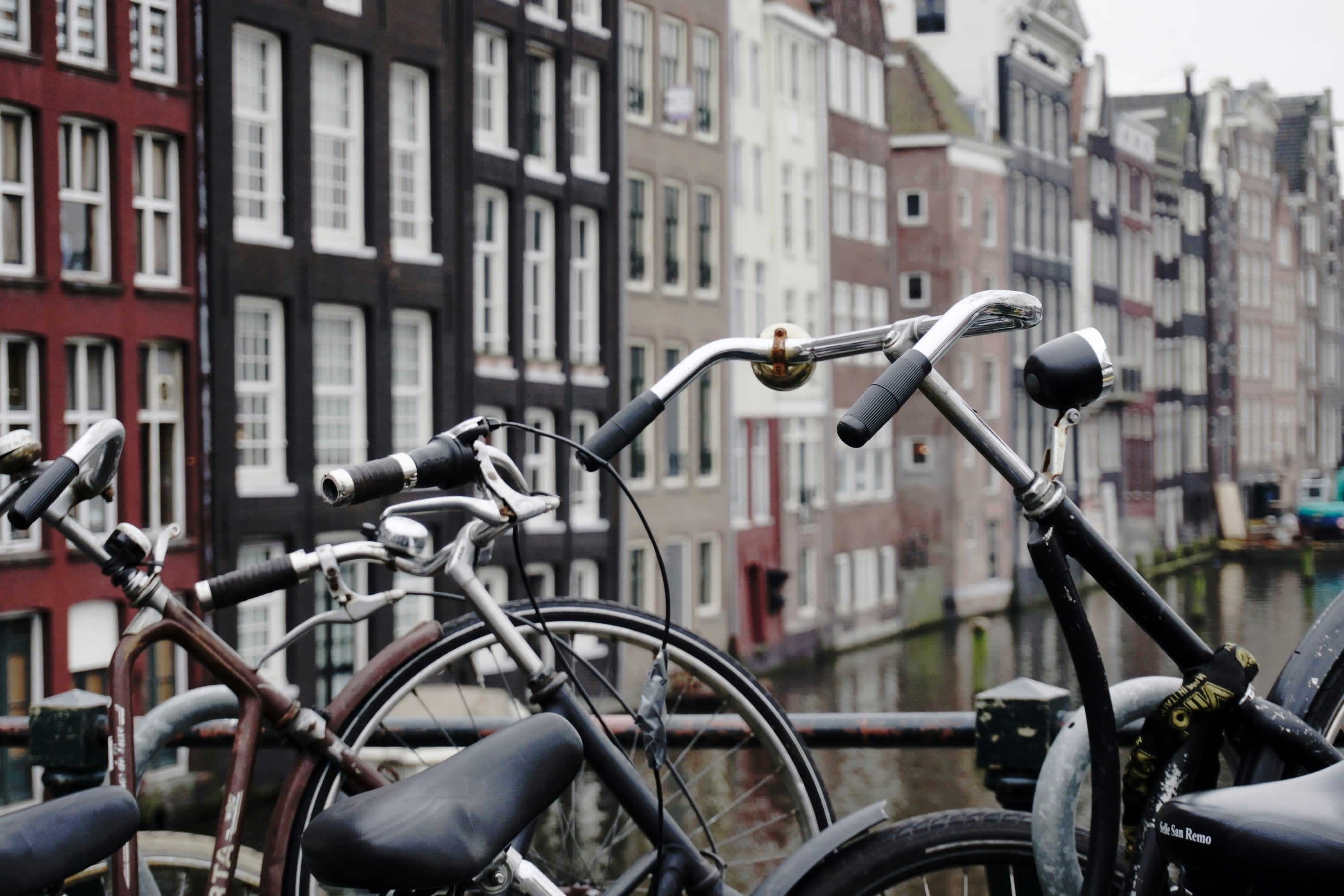 A closeup view of the handlebars of two bicycles that are seemingly locked to a railing that overlooks an Amsterdam canal with historic buildings in the background out of focus