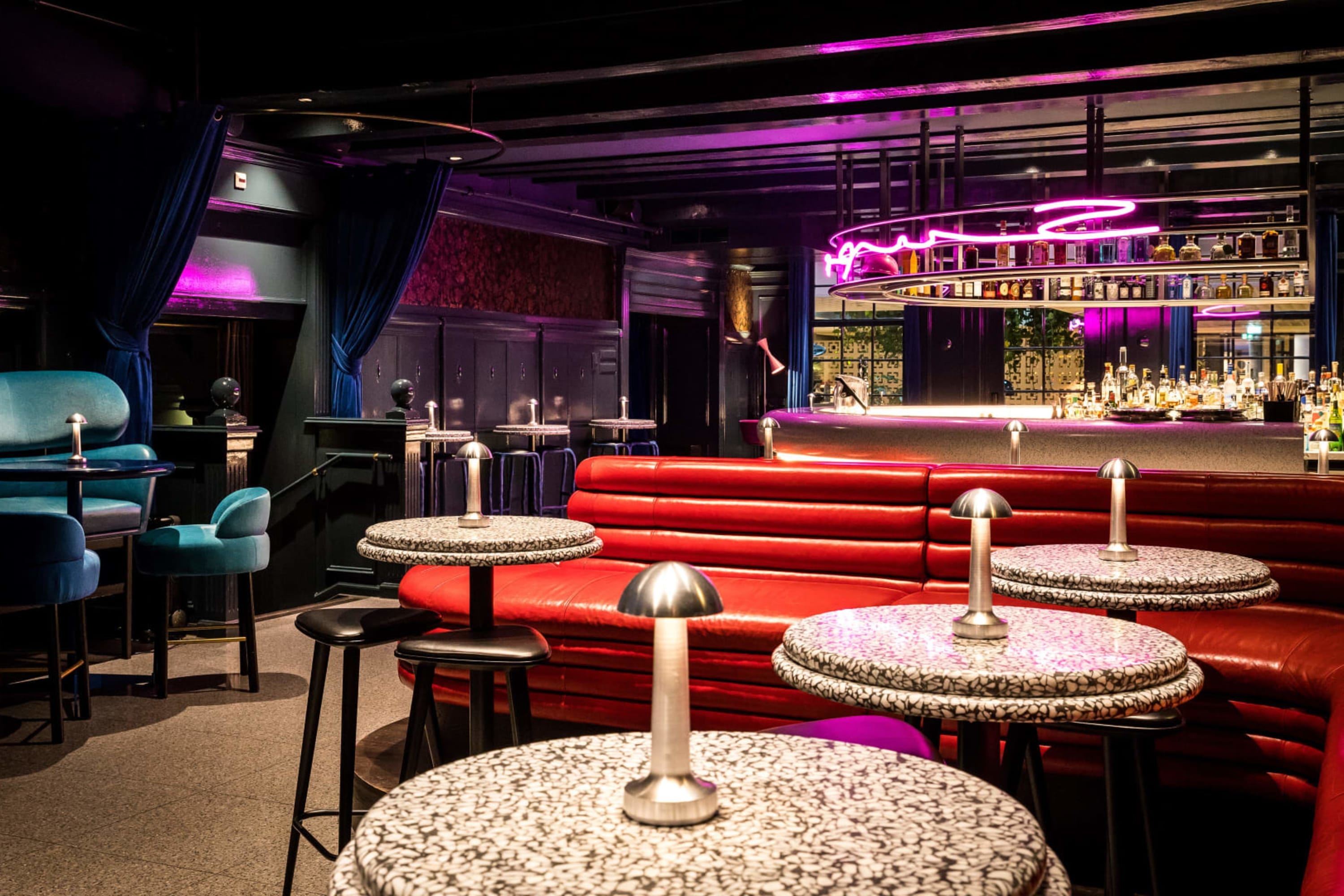 Trendy, retro-esque bar with colorful seating, marble tables and eccentric lighting. The bar is let by a pink neon sign and rows of high-end liquor is visible 