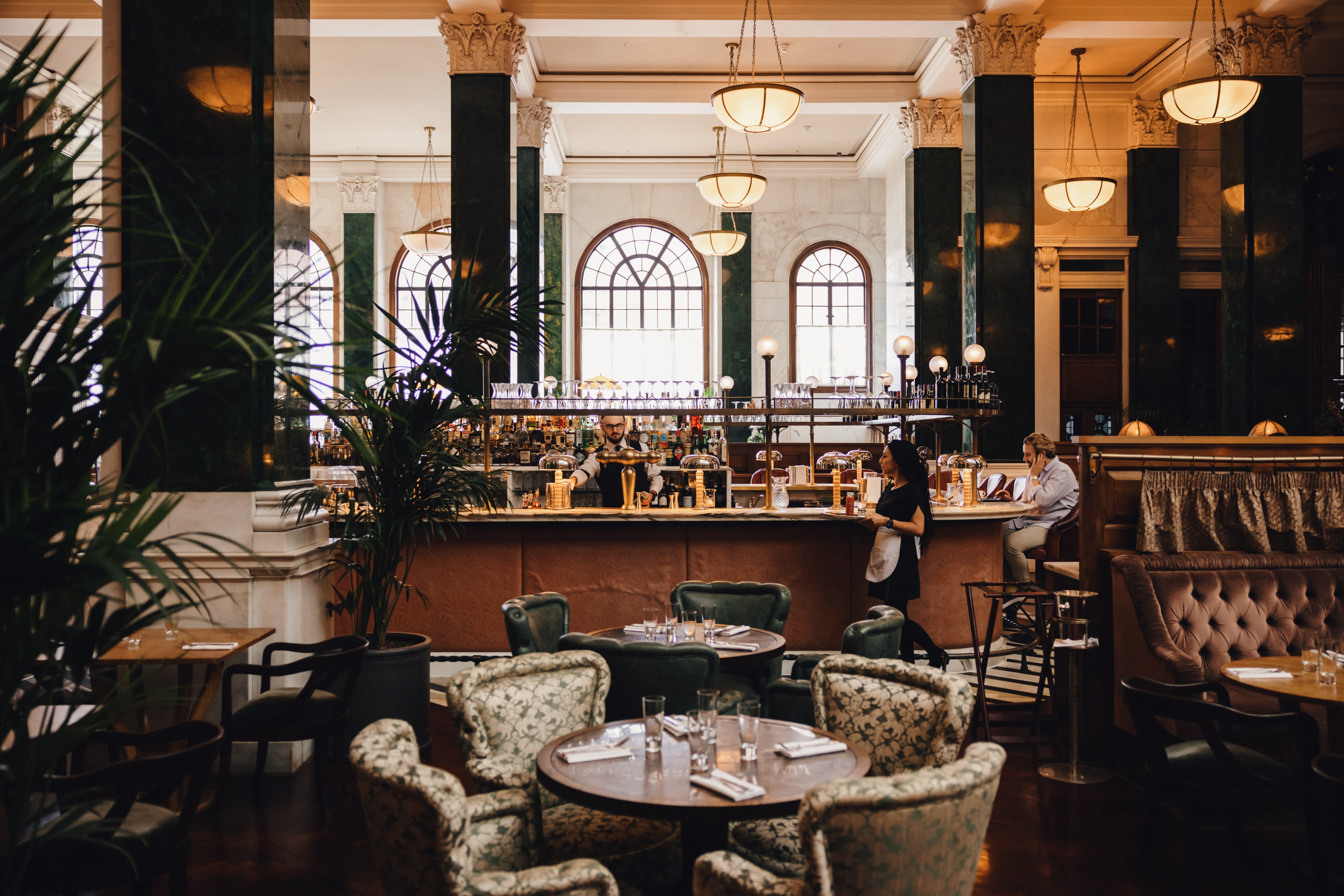 A bar and restaurant area with dark green and brown furnishings and pillars with arched glass windows in view.