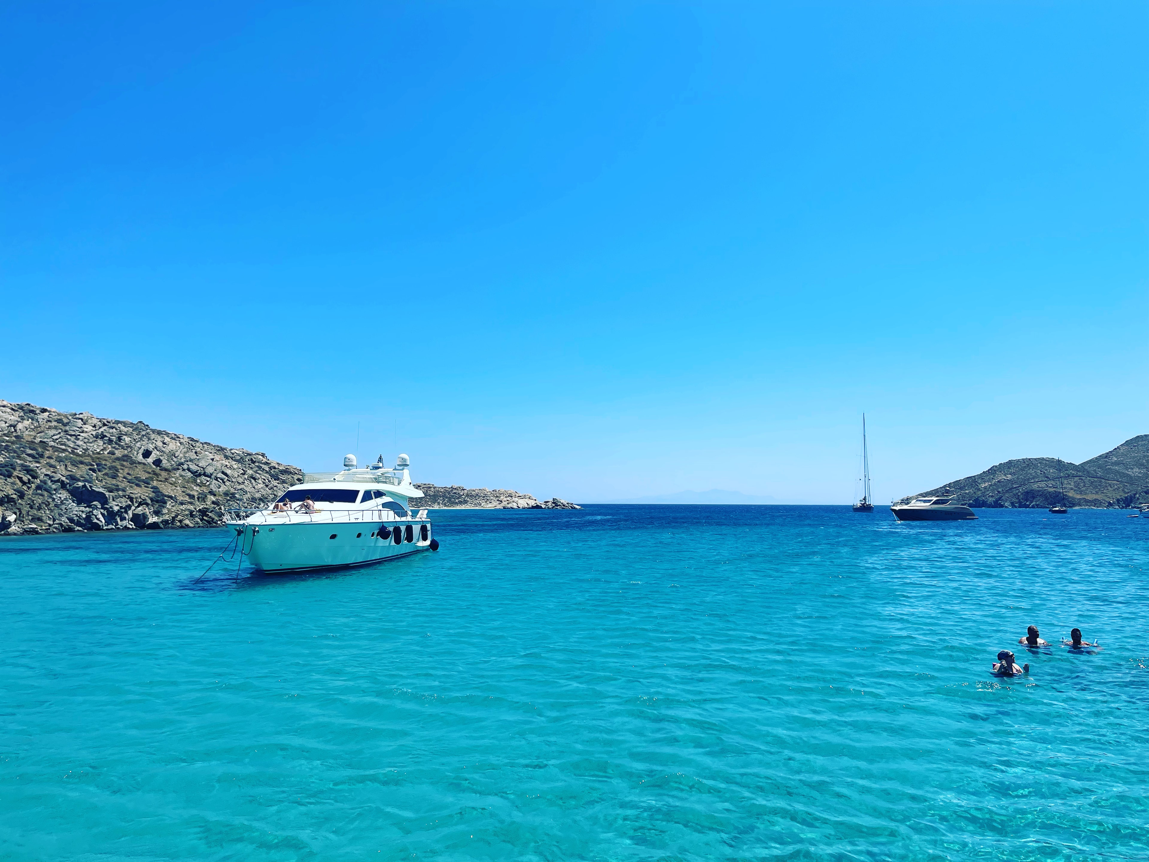 This image depicts a white yacht on top of turquoise blue waters with people floating in the sea on the right side. 