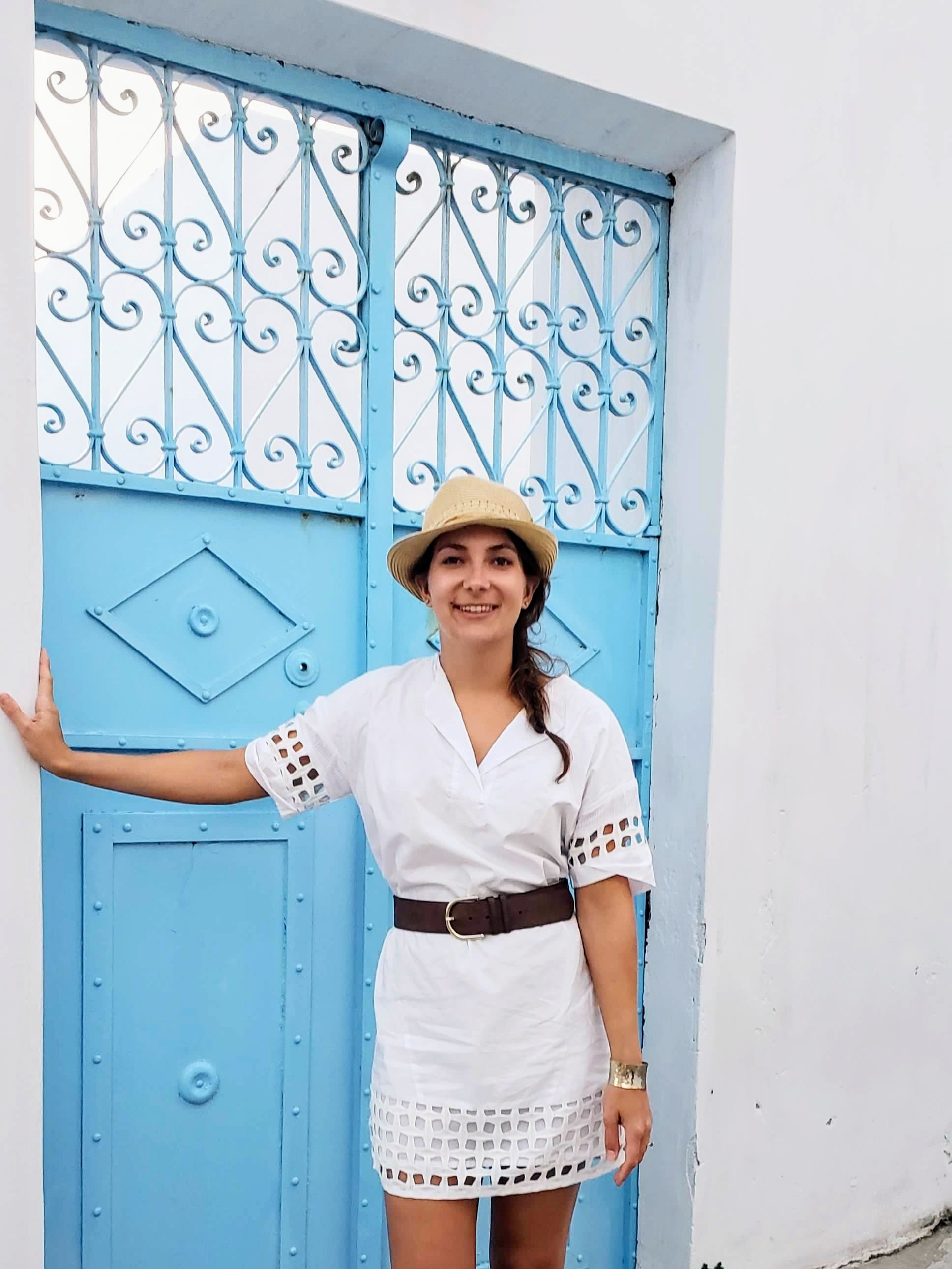 Advisor posing outside in a white outfit with a blue gate in the distance on a sunny day.