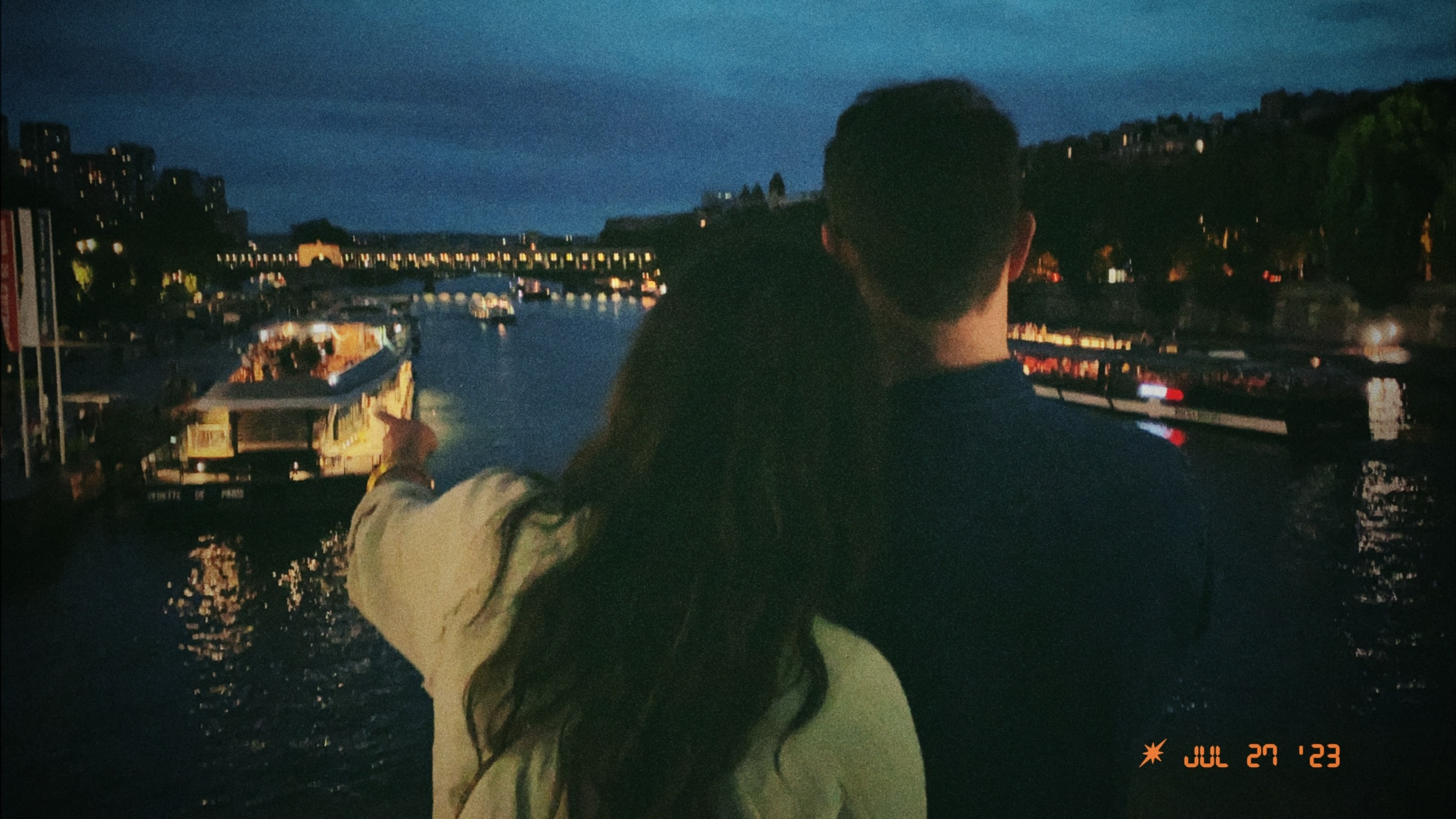 A couple gazing at the ocean with a boat dock lit up at night.