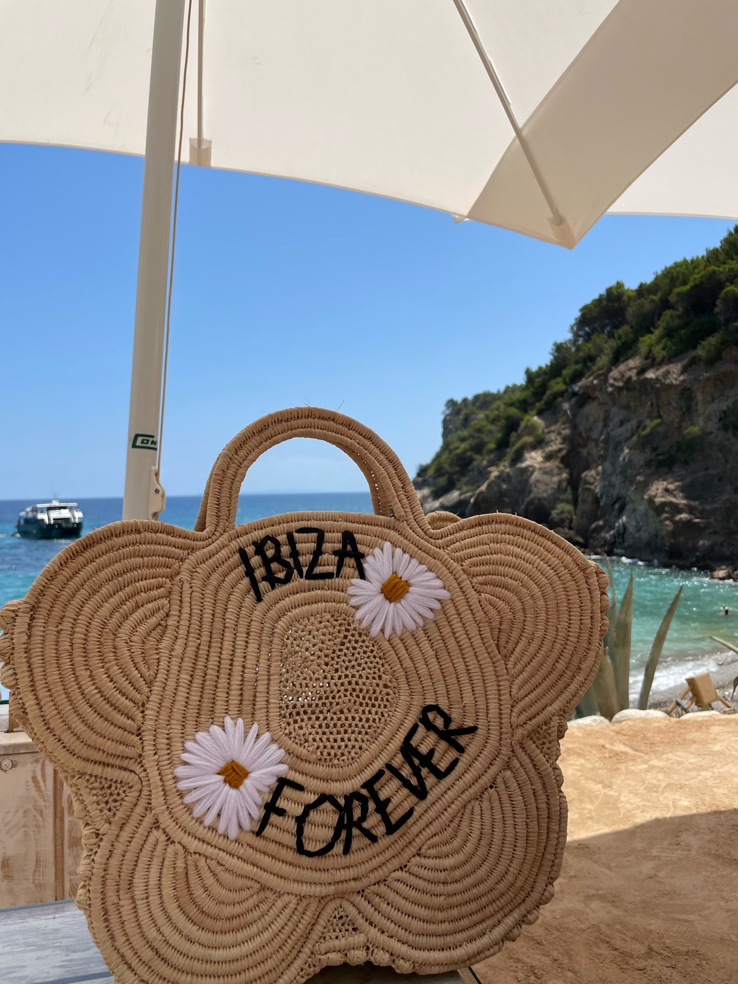 A straw purse with the embroidered text IBIZA FOREVER with a beach view behind.