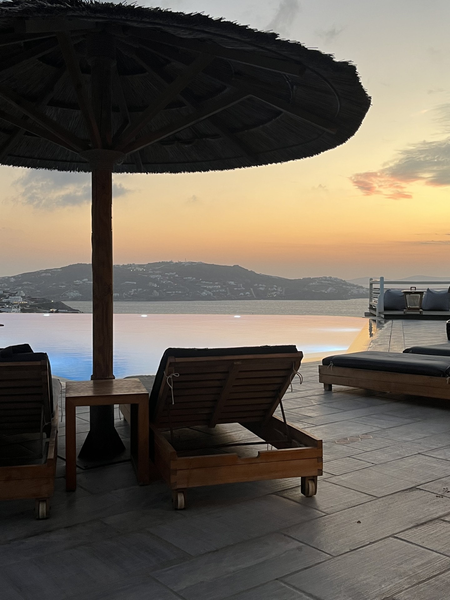 A lounge chair under an umbrella overlooking the water at sunset.