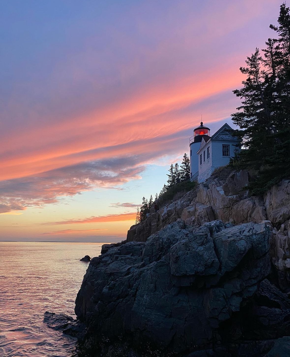 An image of an ocean cliff at sunset with a light house at the edge of the rock formation.