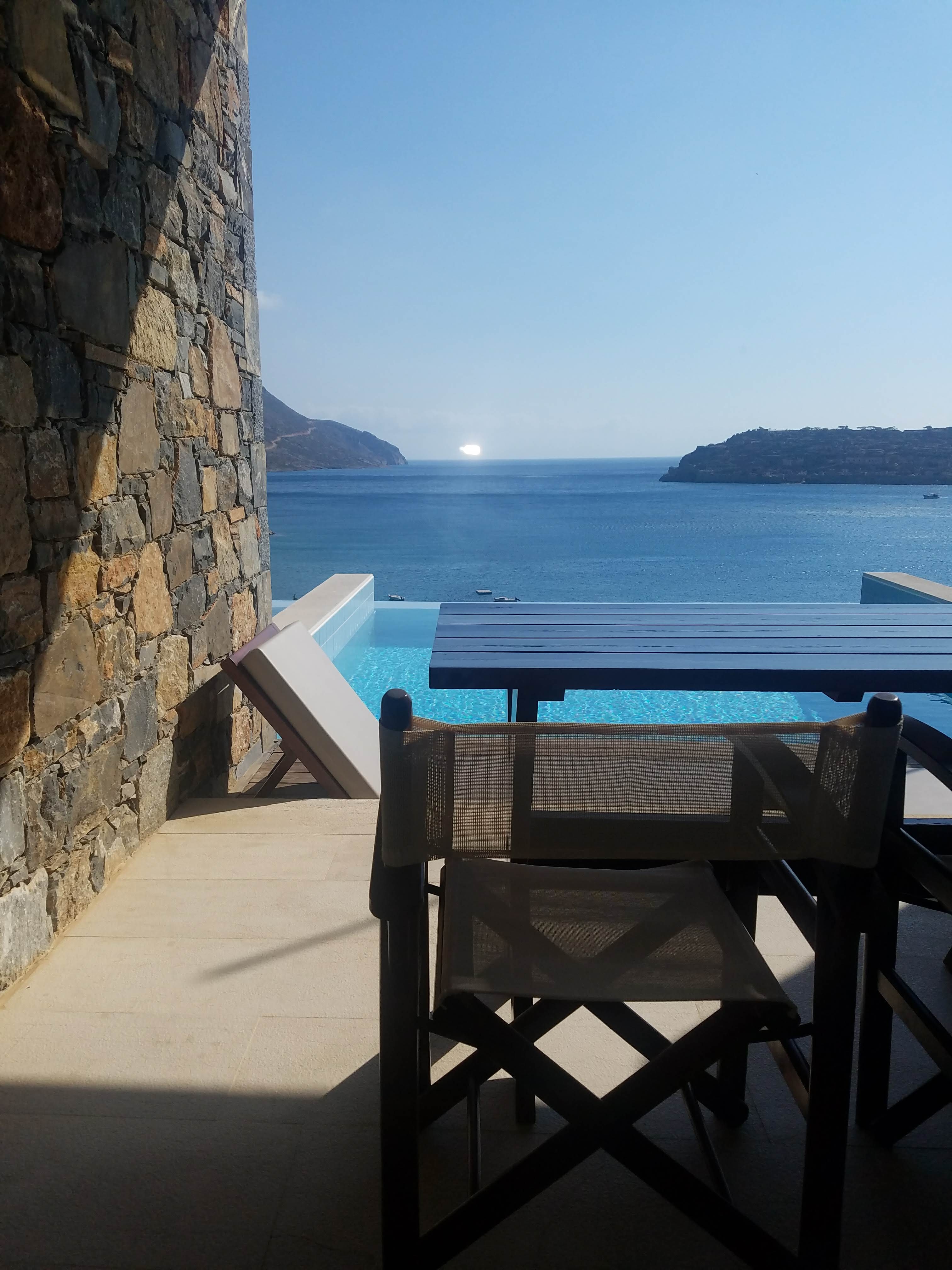 A view of a table and chairs on a balcony with the ocean in the distance on a sunny day.