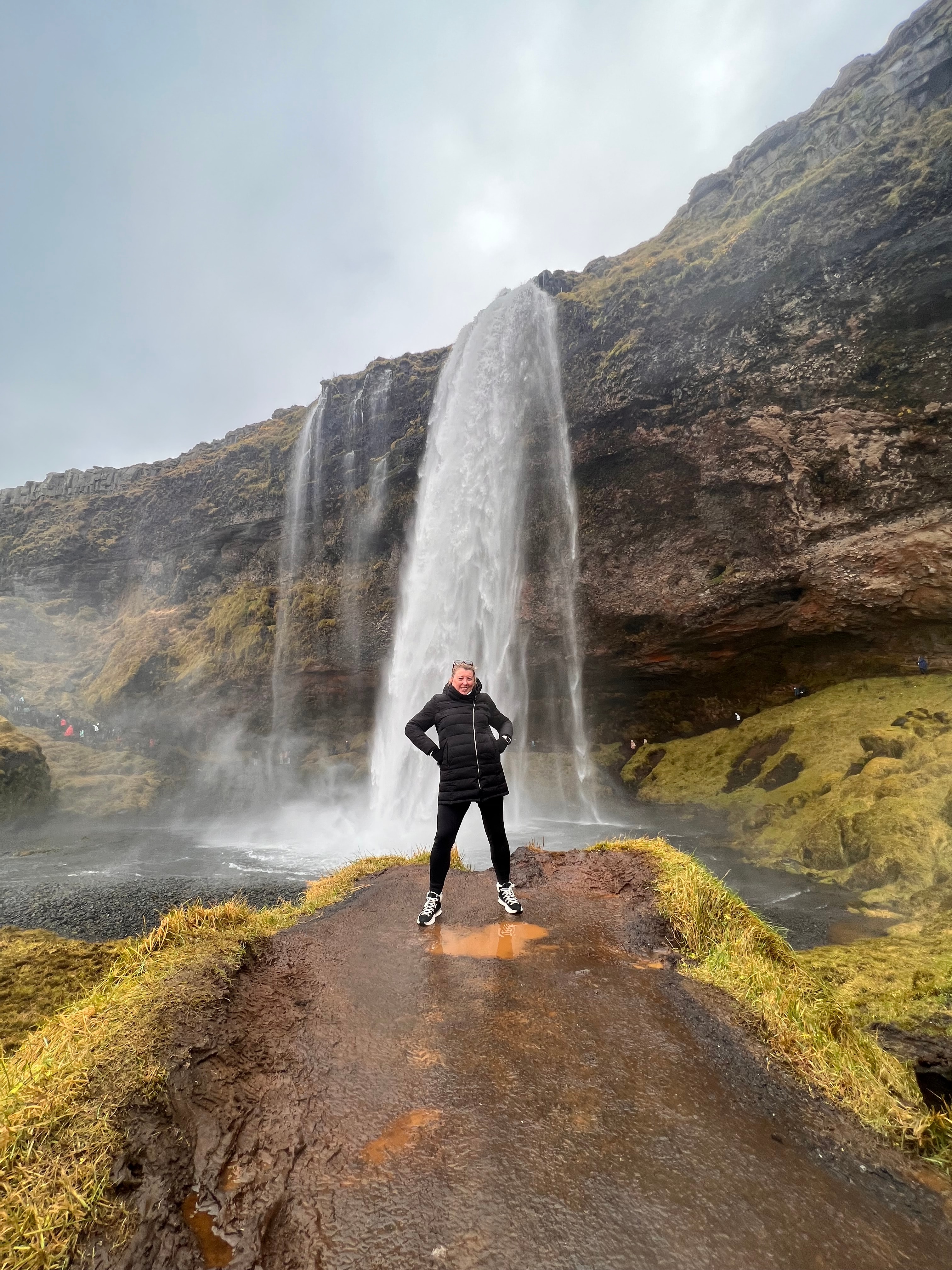 Andrea in all black with her hands on her hips posing in front of a beautiful waterfall on a cloudy day