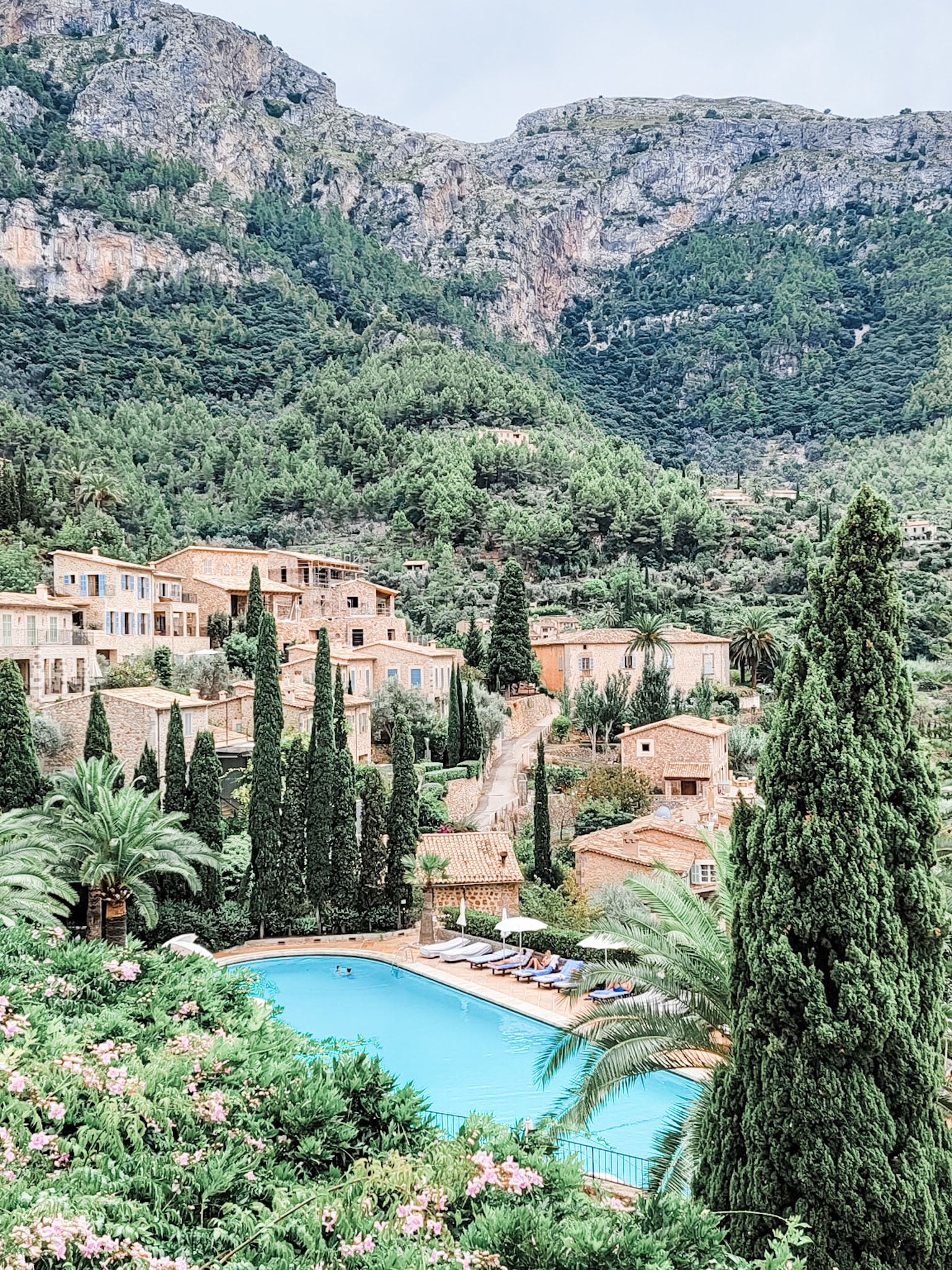 An aerial view of a beautiful pool in a European region with a town in the distance surrounded by foliage and a mountain scape.