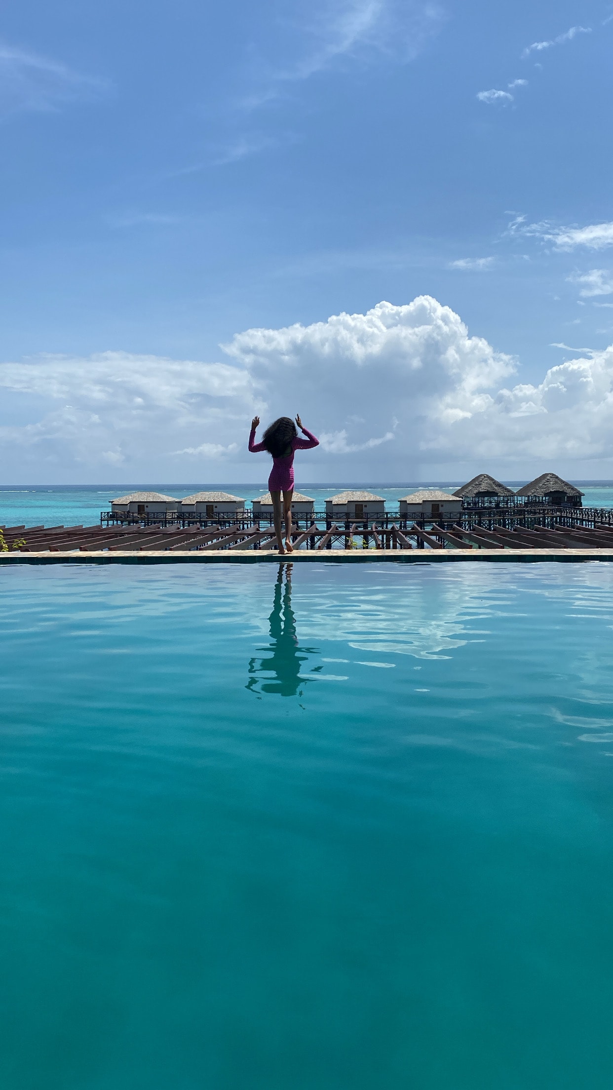 View of Favour standing at the edge of an infinity pool near lounges overlooking the sea on a sunny day