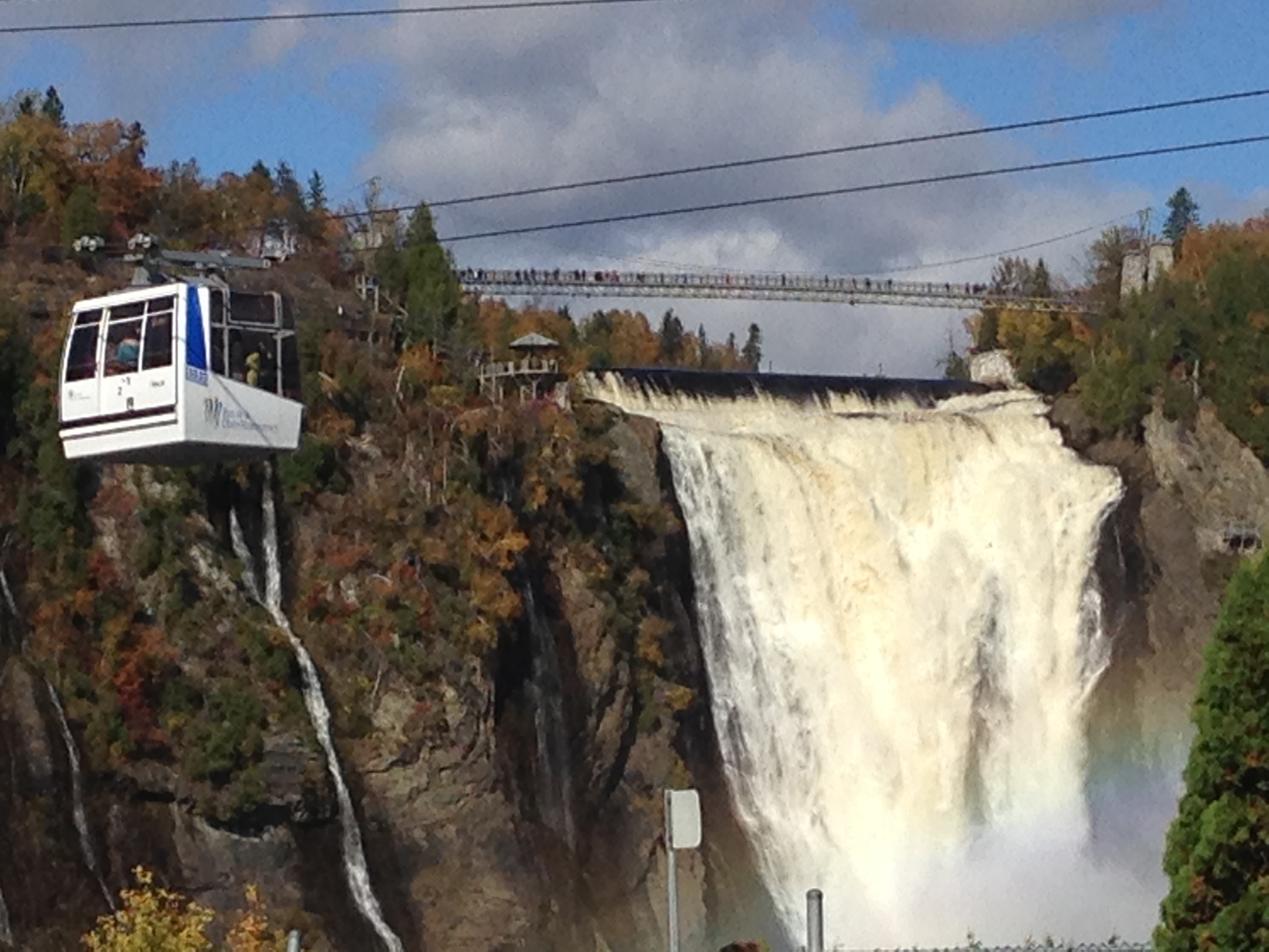 View of a white lift traveling on a cable across from a large cascading waterfall on a sunny day