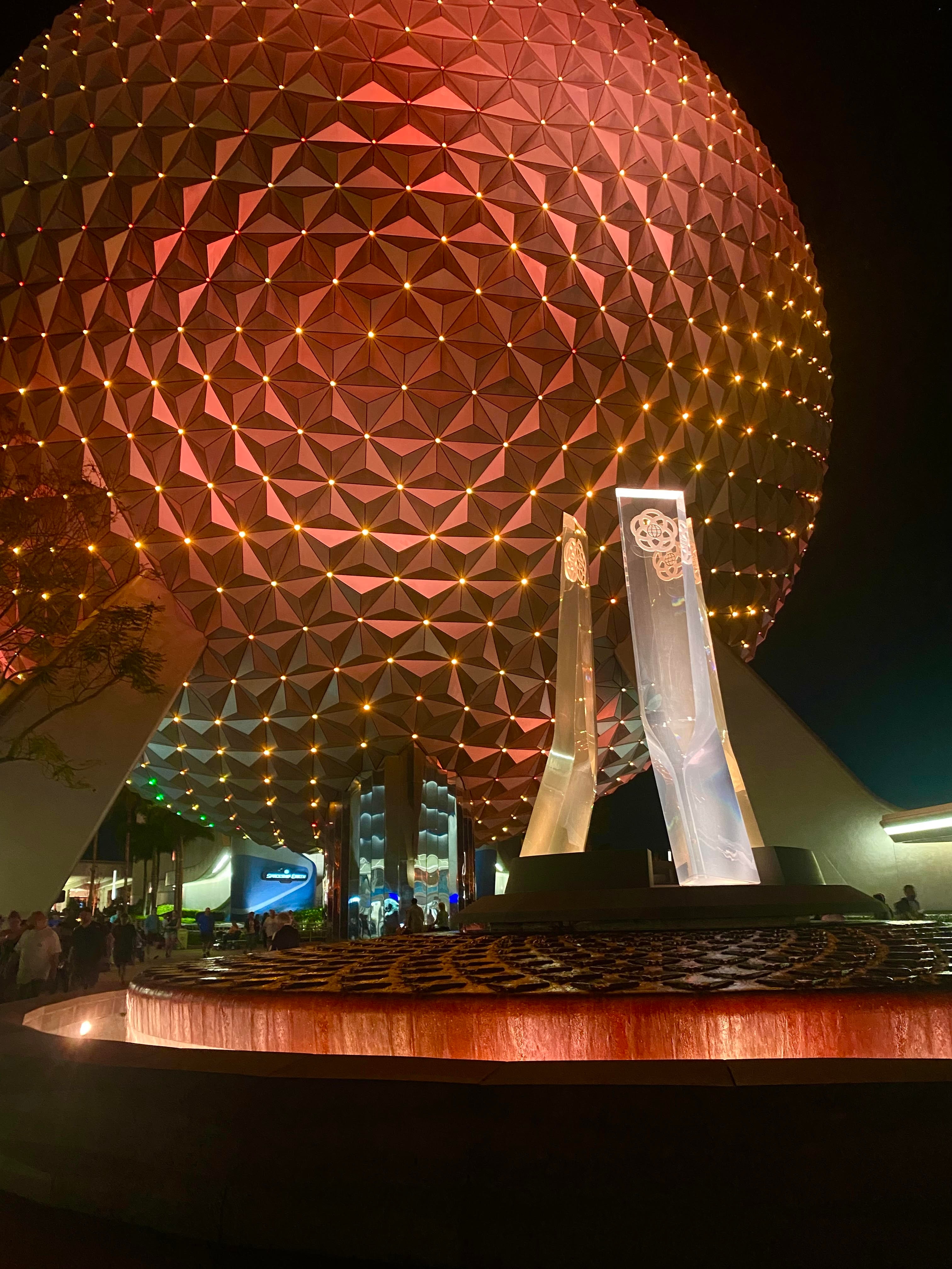 A large circular structure with lights at nighttime with people walking in the distance. 