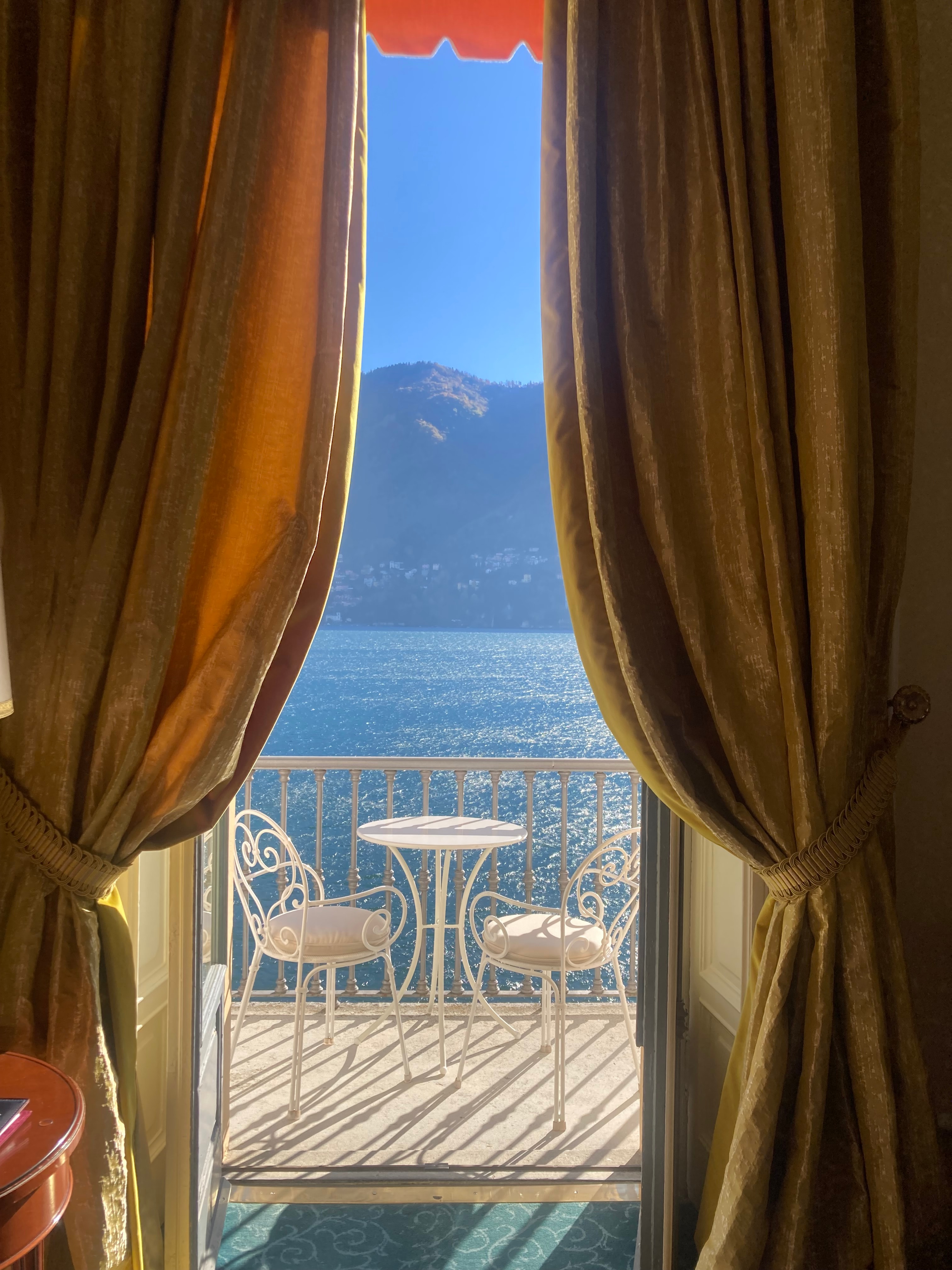 A view of a hotel balcony with luxurious curtains and an iron outdoor table a chairs with the ocean and a mountain range in the distance.