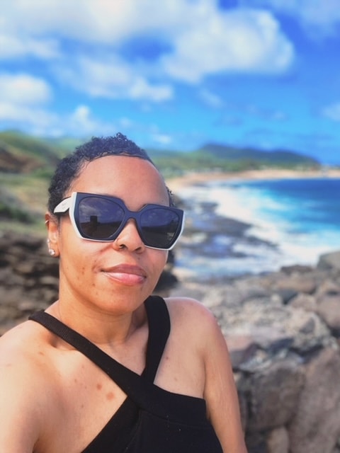 Dominique taking a selfie in big black sunglasses with a beautiful coastline visible behind her on a sunny day