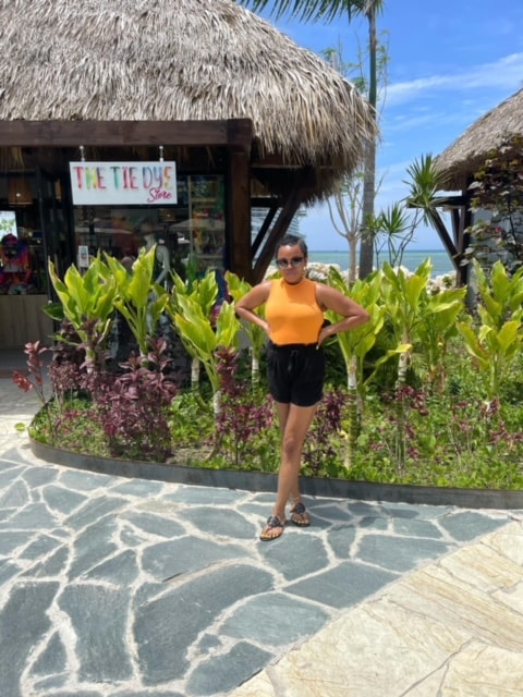 Dominique in an orange shirt and black shorts posing in front of plants and buildings with thatched rooftops on a sunny day