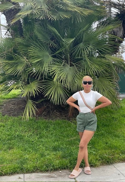 Dominique in a white shirt and green shorts and hands on her hips posing in front of a large palm tree