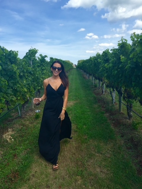 Whitney in a black dressing holding a glass of wine standing in between trees in a vineyard