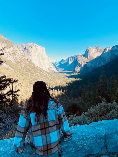 View of Whitney in a plaid jacket sitting on a rock facing a beautiful mountain landscape on a sunny day