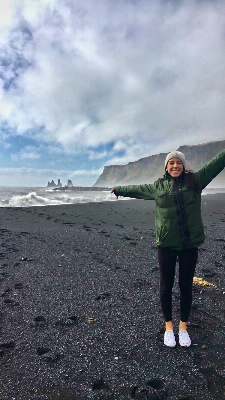 Whitney in winter clothing posing on black sand with snowy mountains in the distance