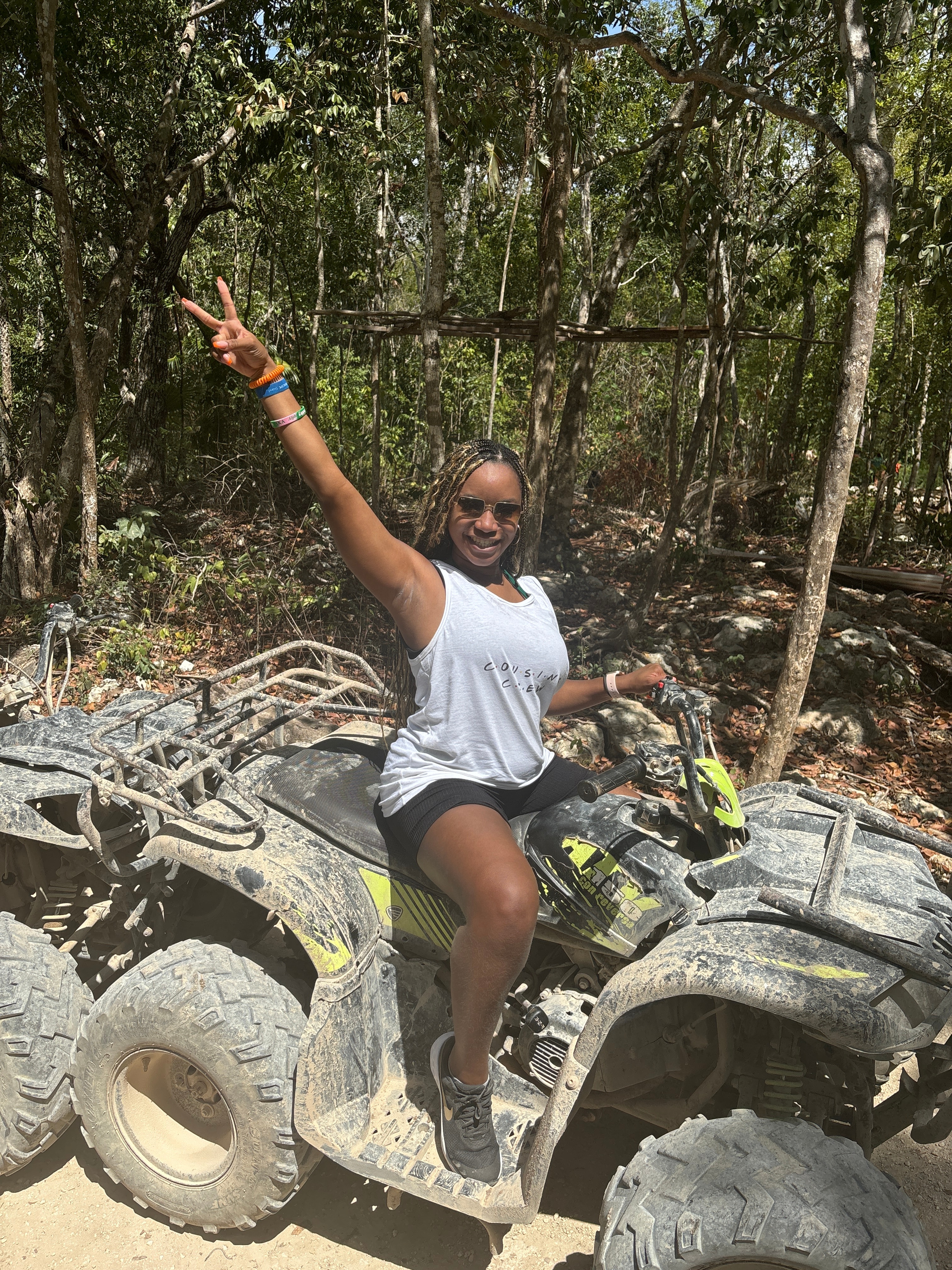 Jazmyn in a white tank top and arm raised sitting on a dirty ATV with jungle visible behind her