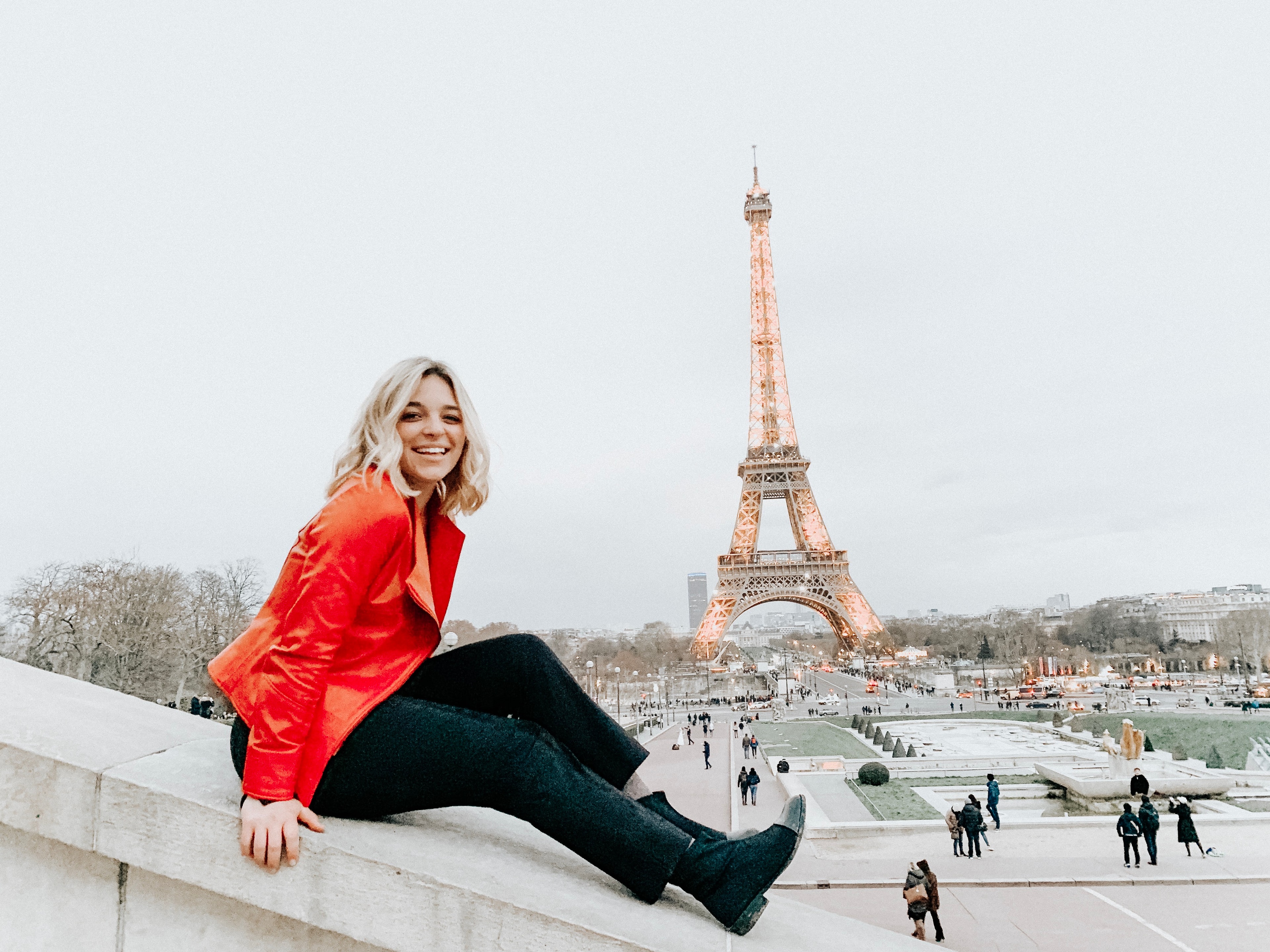 Hannah in bright red jacket sitting on a ledge with a beautiful view of the Eiffel Tower behind her